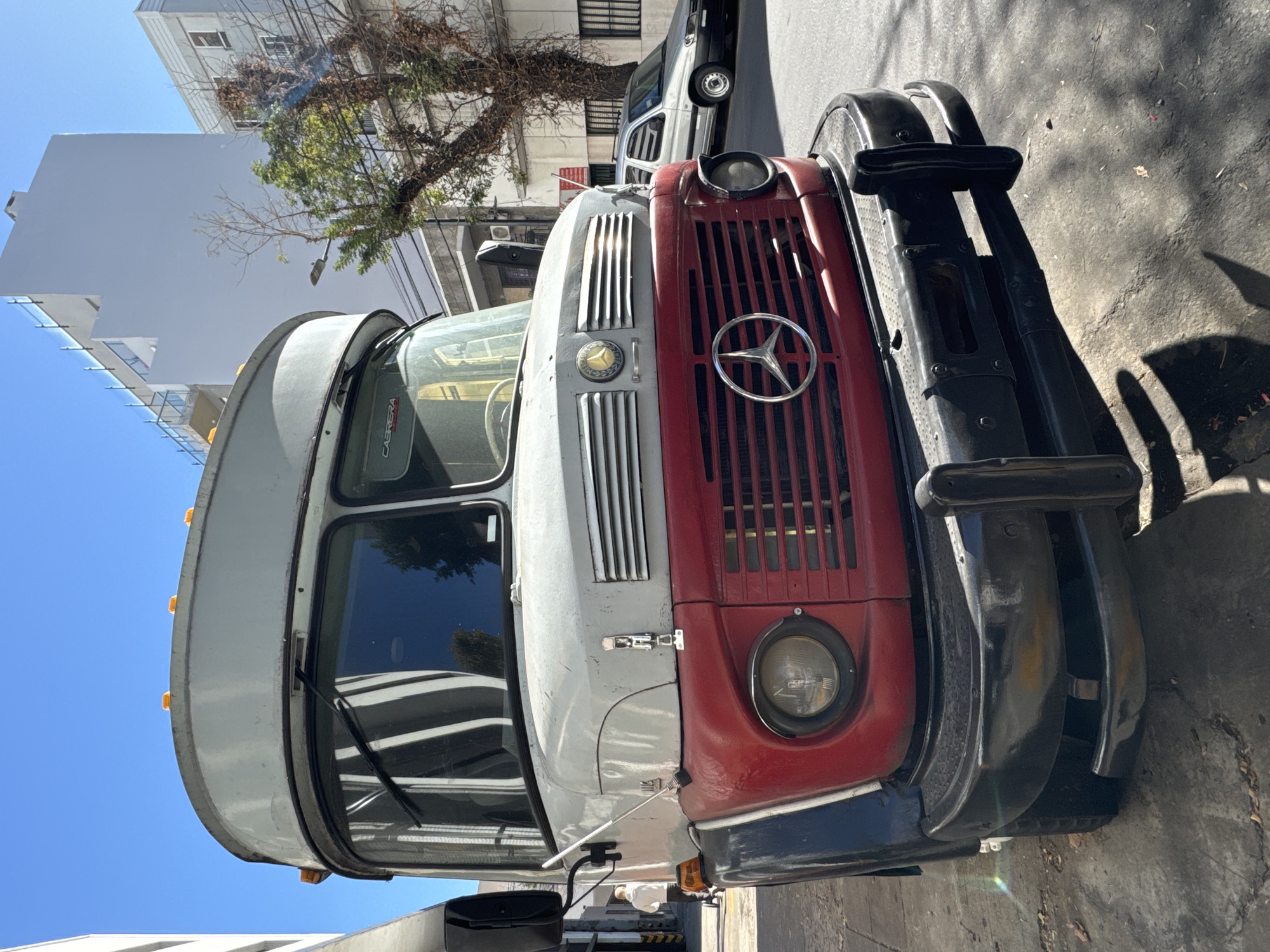 A vintage Mercedes-Benz bus is parked on a street in Buenos Aires, Argentina. The bus features a red and gray color scheme with a prominent Mercedes-Benz star emblem on the red grill. The front bumper is black. The background includes a white building under a blue sky, and a tree partially obscures another building. A modern white van is also parked nearby. The scene is captured during daylight hours with shadows cast on the pavement.