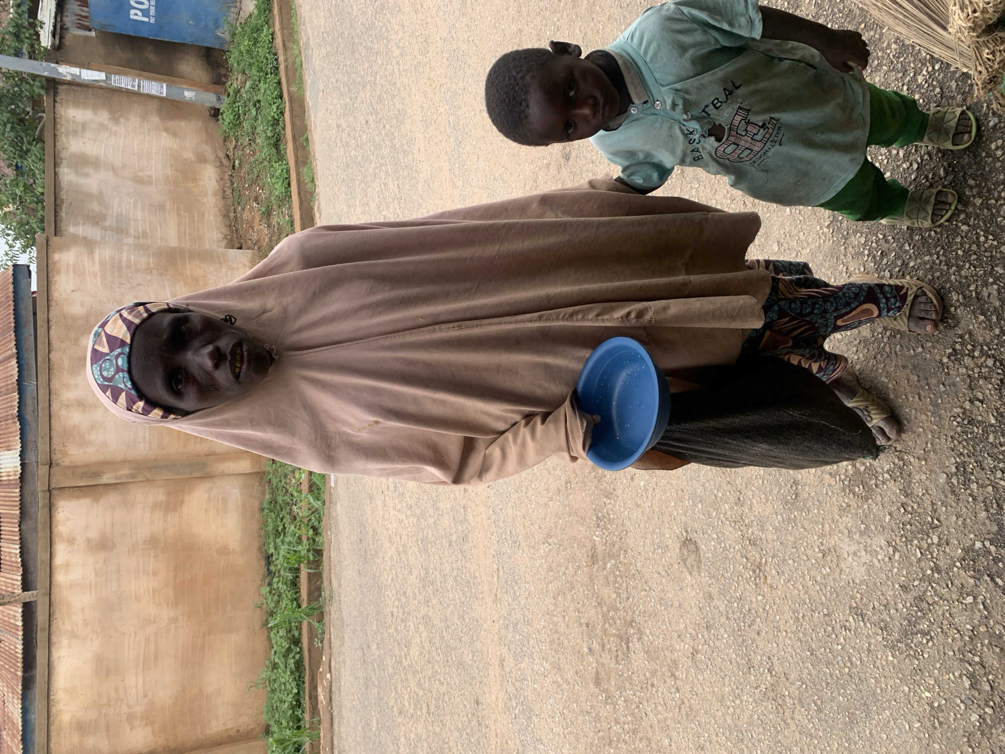 An elderly, thin, dark-skinned woman wearing a tan headscarf and a dark skirt stands on a paved surface. She holds a blue plastic bowl in her left hand, seemingly soliciting something. To her right, a young dark-skinned boy in a light blue shirt and green pants stands. They are likely in Jos, Nigeria, judging from the location metadata. Behind the woman is a wall with vegetation growing along its base. A corrugated metal structure is visible to the left of the wall. A blue sign is partially visible in the upper left corner. The pavement appears cracked and uneven in some areas.