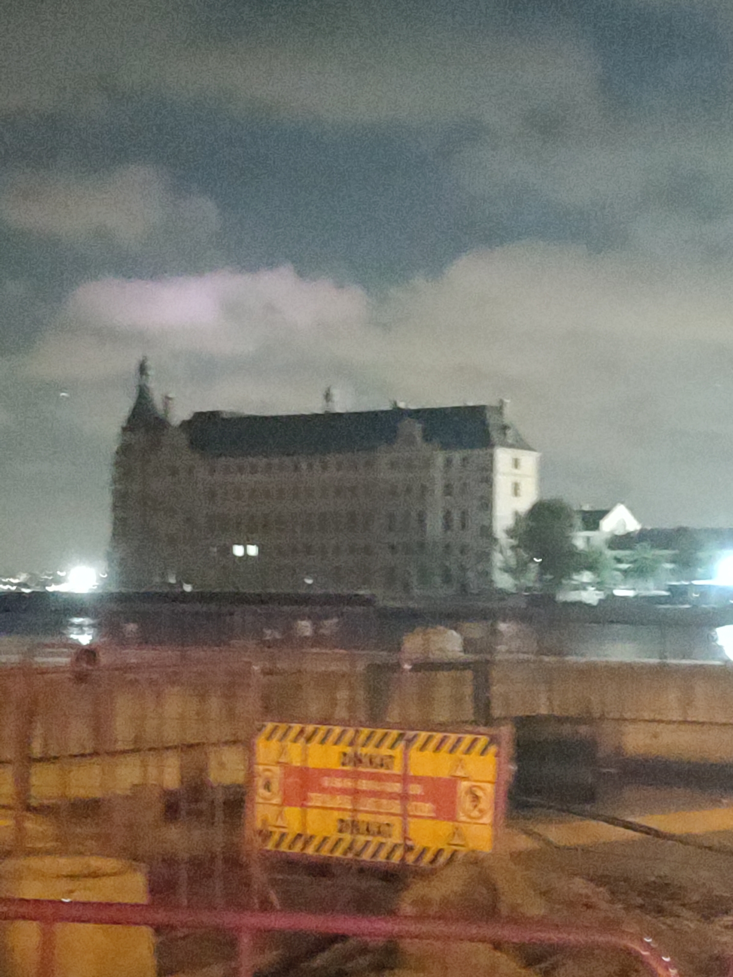 The image depicts a nighttime scene in Kadıköy, Turkey, featuring the historic Haydarpaşa Train Station. The station, a large, multi-story building with a distinctive tower, is partially illuminated, with lights visible in some of its windows. The sky above is cloudy and dark, with some faint light sources suggesting urban illumination. In the foreground, there is a construction or barrier area with a yellow and black striped warning sign. This sign appears to have text and hazard symbols, indicating potential danger. The overall image is slightly blurred, contributing to a somewhat hazy and atmospheric depiction of the location at night.