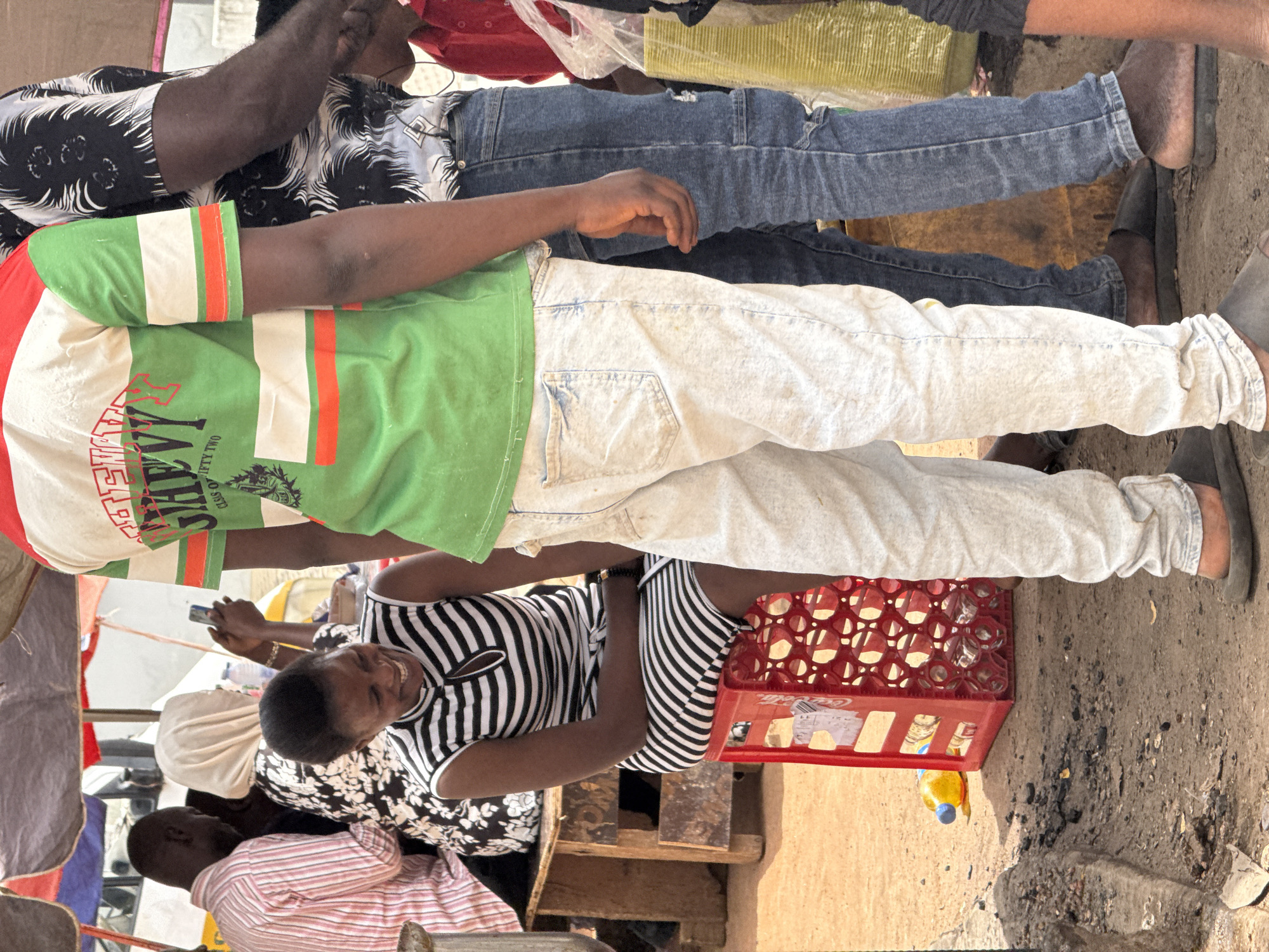 Several individuals are gathered in what appears to be an outdoor marketplace or street scene, possibly in Abuja, Nigeria, given the context. A woman wearing a black and white striped top is looking at a phone with a smile. There is a red Coca-Cola crate containing glass bottles nearby. The ground is dirt and gravel. Other individuals are visible, some wearing patterned shirts and jeans. The scene seems to capture a moment of interaction and commerce.