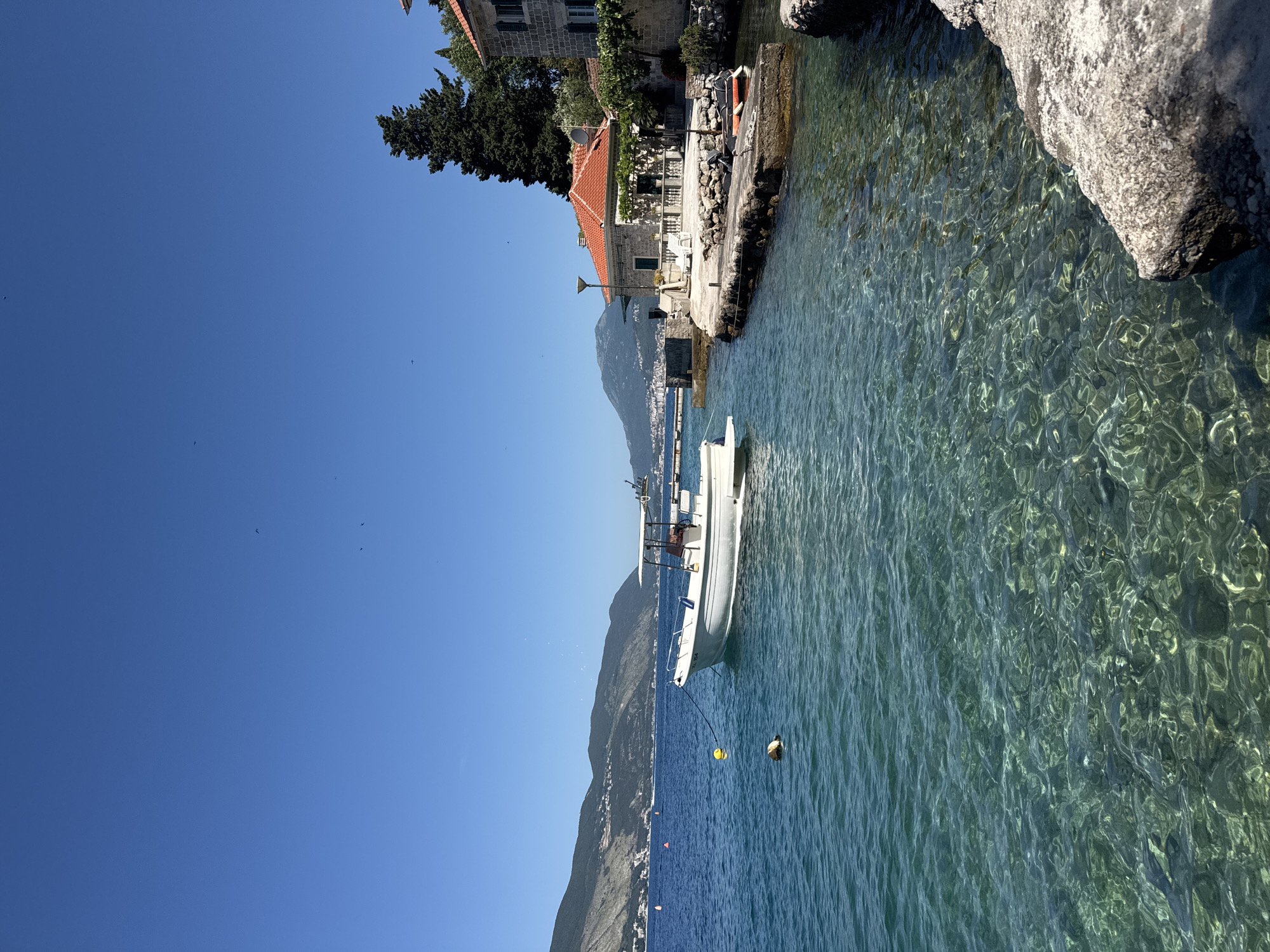 The image displays a bright, sunlit coastal landscape in the city of Unknown, Unknown. The foreground is dominated by exceptionally clear, calm turquoise water, which reveals the rocky seabed with distinct light patterns from the sun. A white motorboat with a canopy is moored slightly left of center, attached by a black rope to a yellow spherical buoy. A smaller white and beige cylindrical buoy floats nearby.
Further into the midground, the water deepens to a rich blue, stretching towards a distant shoreline. This distant shore is backed by a range of green and rocky mountains, upon whose slopes scattered buildings indicate a settlement.
Along the right side of the image, traditional stone buildings with red tiled roofs line the immediate shore. One prominent two-story stone building features a white balustrade patio area with a single white lounge chair, a green door, and a satellite dish mounted on its red roof. A dark-colored street lamp stands in front of this building. Behind and between the structures, several dark green coniferous trees, possibly cypresses, are visible. Adjacent to the stone building, a small red inflatable dinghy with a white outboard motor is moored next to a concrete jetty.
The sky is a clear, cloudless deep blue, occupying the upper portion of the frame. Several small, dark, V-shaped specks, likely birds, are visible scattered across the vast expanse of the sky. No people are discernible in the scene.