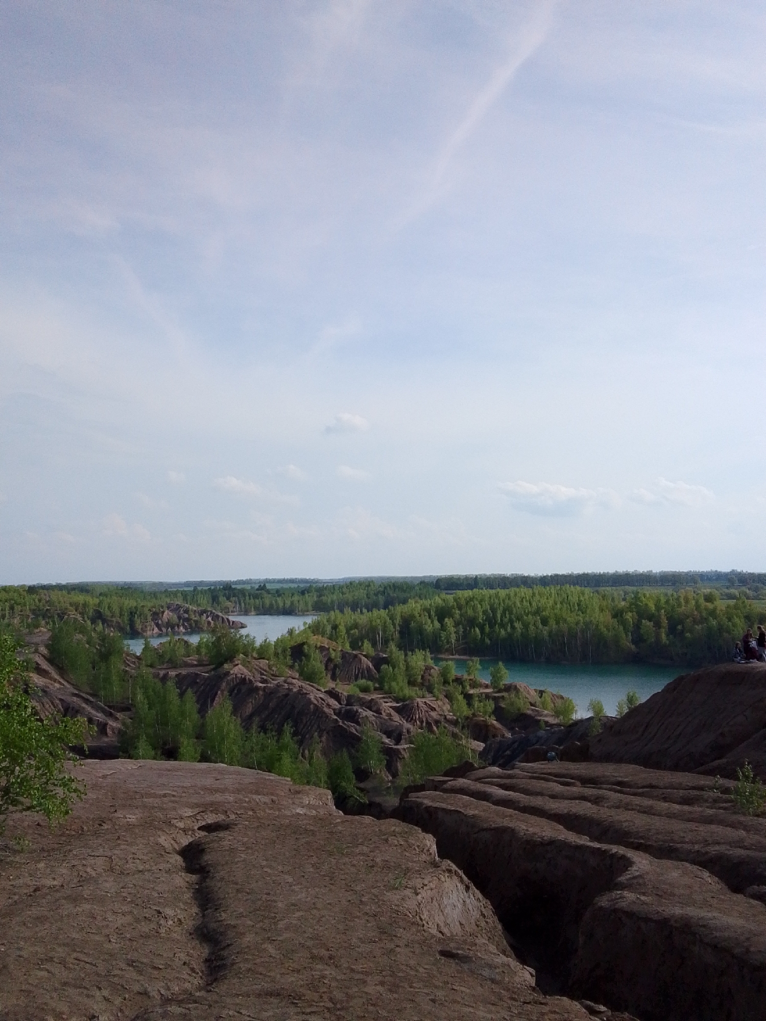 The image depicts a landscape featuring a body of water surrounded by forested areas and undulating terrain. The terrain is characterized by ridges and valleys, suggesting a quarry or mining area. The water is a light turquoise color. The sky is partly cloudy. In the distance, there are indistinct figures of people. The location is in the city of Unknown, Unknown.