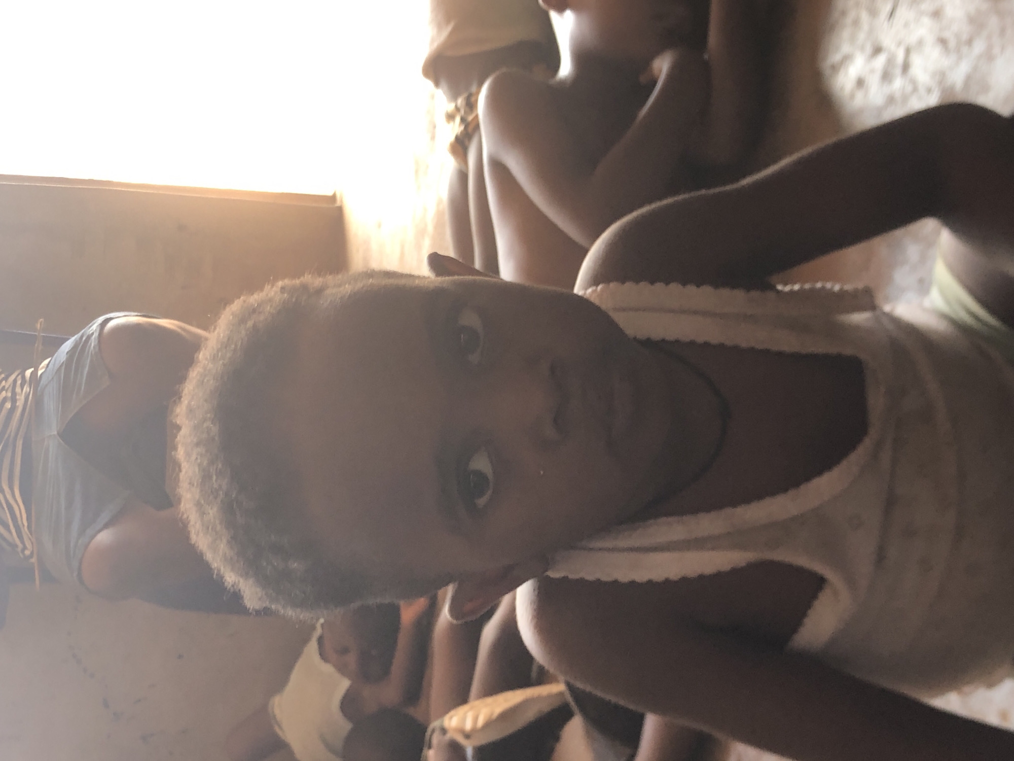 In a building in Ukwu-Abwa, Nigeria, a child with curly hair and wearing a white tank top looks directly at the camera. Several other children and an adult wearing a striped shirt are visible in the background. The environment appears dimly lit with natural light entering from an unseen window.