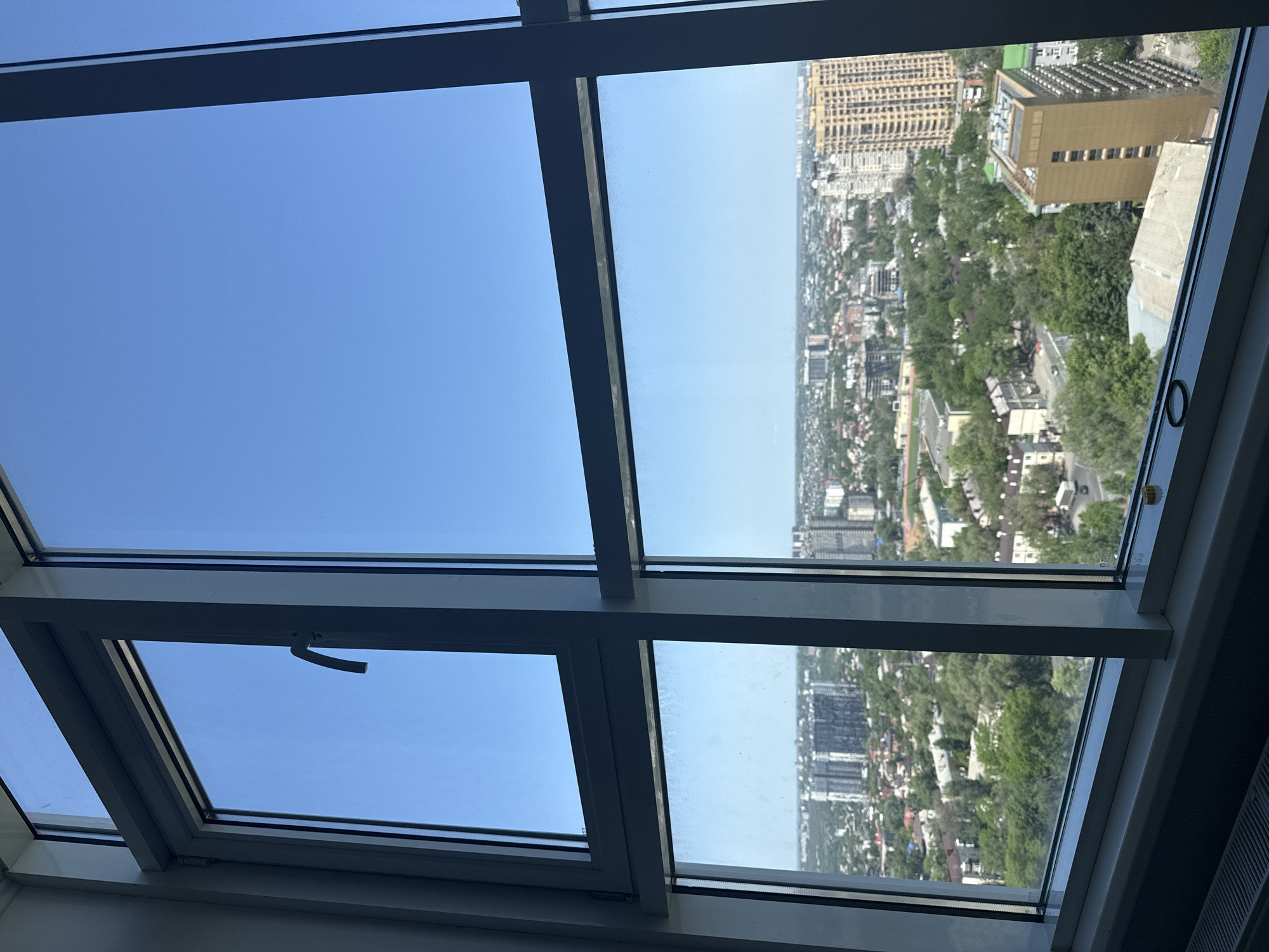 The image is a view from a high vantage point, likely an apartment or office building, in what appears to be a sunny Rostov-on-Don, Russia. The scene is framed by a modern window with a metal frame, divided into multiple panes. The left panes show a clear, blue sky. The right panes offer a panoramic view of the city. The cityscape includes a mix of high-rise buildings, lower residential structures, and a lot of green spaces that indicate trees. The architecture in the distance seems to be mixed, with both older and modern buildings visible.