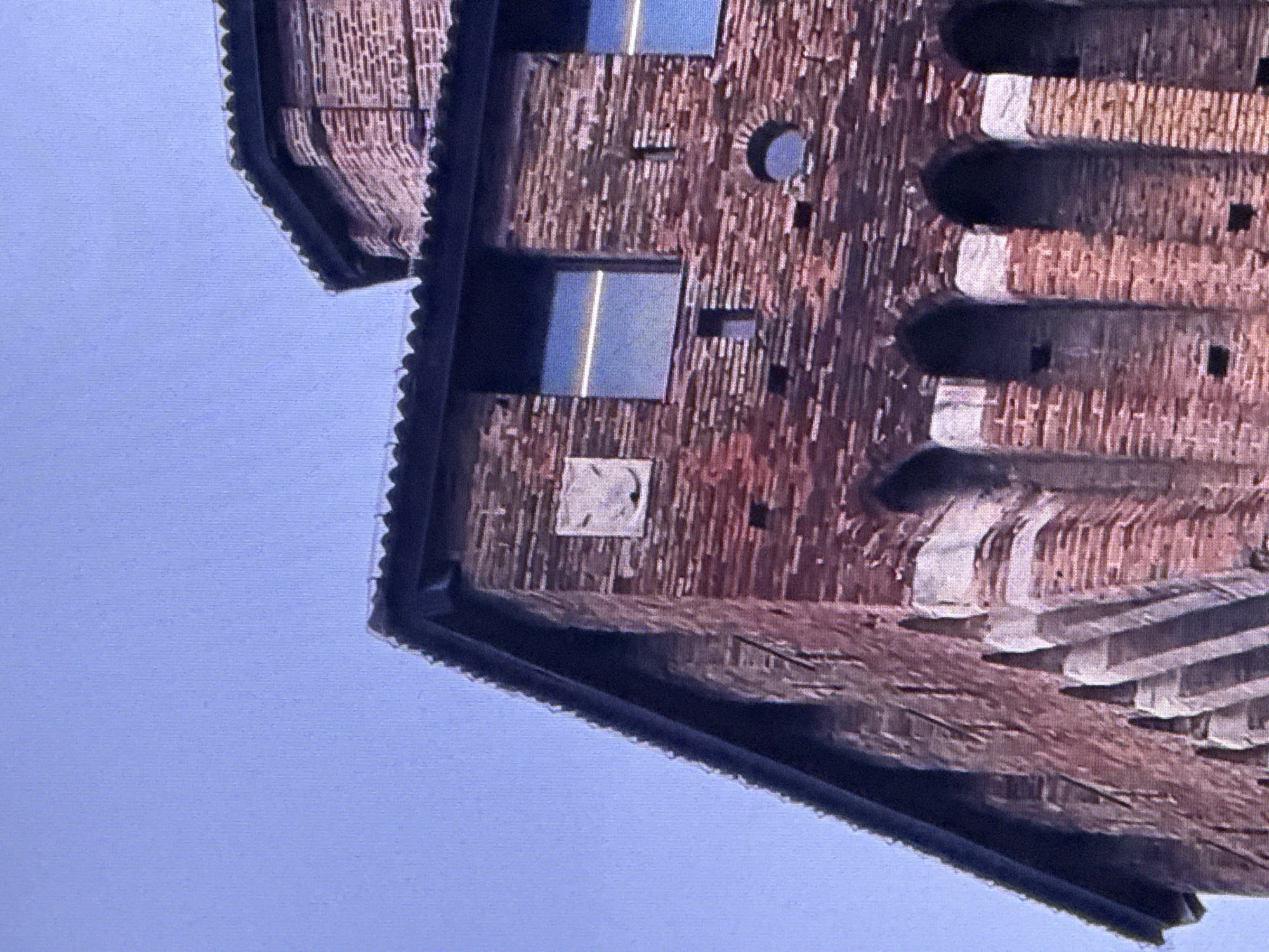 The image shows the upper section of a red brick building against a clear blue sky. The building has a tiled roof and features architectural elements such as arched openings and rectangular windows. A small, light-colored emblem with a stylized face is visible on one of the brick sections. The overall impression is of an older, possibly historical, structure. There are no people or animals visible in the image, and the focus is solely on the architecture of the building. The image does not depict any recognizable landmarks or features of the city of Dubai, United Arab Emirates.