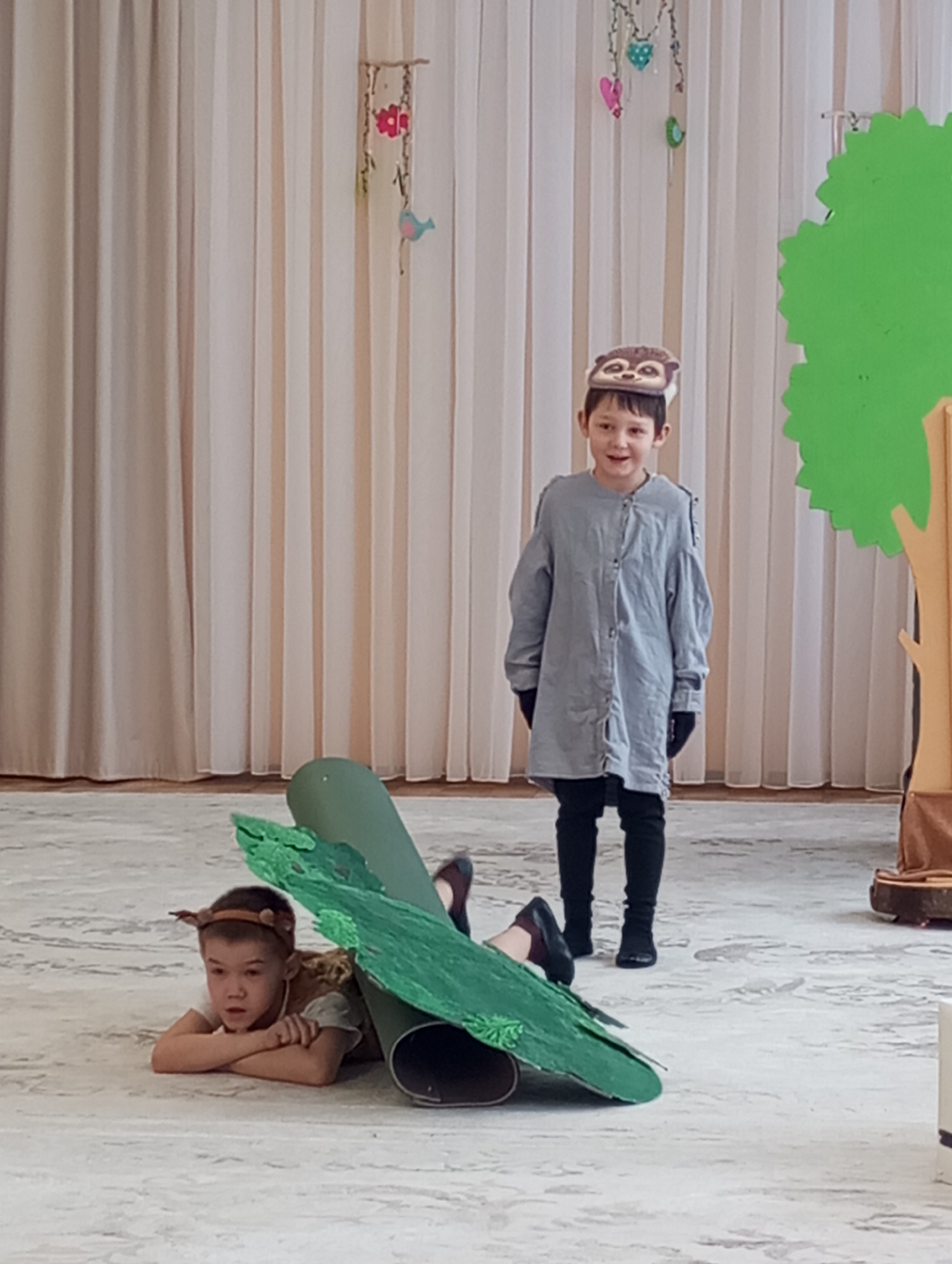 Two young boys are performing in what appears to be a children's play or theatrical event, likely in a school or kindergarten in Izhevsk, Russia. The indoor setting features light-colored, sheer curtains as a backdrop, with decorative elements hanging from a stick, including red flowers, pink hearts, a blue bird, and green leaf shapes. A light-colored patterned floor covers the performance area.
On the right side of the frame, a simple stage prop of a tree with a brown trunk and a bright green, cut-out leafy canopy stands. One boy, dressed in a grey long-sleeved tunic, black pants, and black gloves, stands upright, wearing an animal-themed hat resembling a badger or similar creature. He is smiling and looking towards the viewer, seemingly engaged in his role.
Closer to the foreground, another boy is lying on his stomach, propped up on his elbows, wearing a light-colored shirt and a headband with animal ears, possibly depicting a deer. He has a focused or neutral expression. A large, green prop, possibly representing a rolled-up log or oversized leaf with smaller green foliage attached, is positioned next to and partially covers him. The overall scene suggests a creative and imaginative children's performance. There is no visible text in the image.