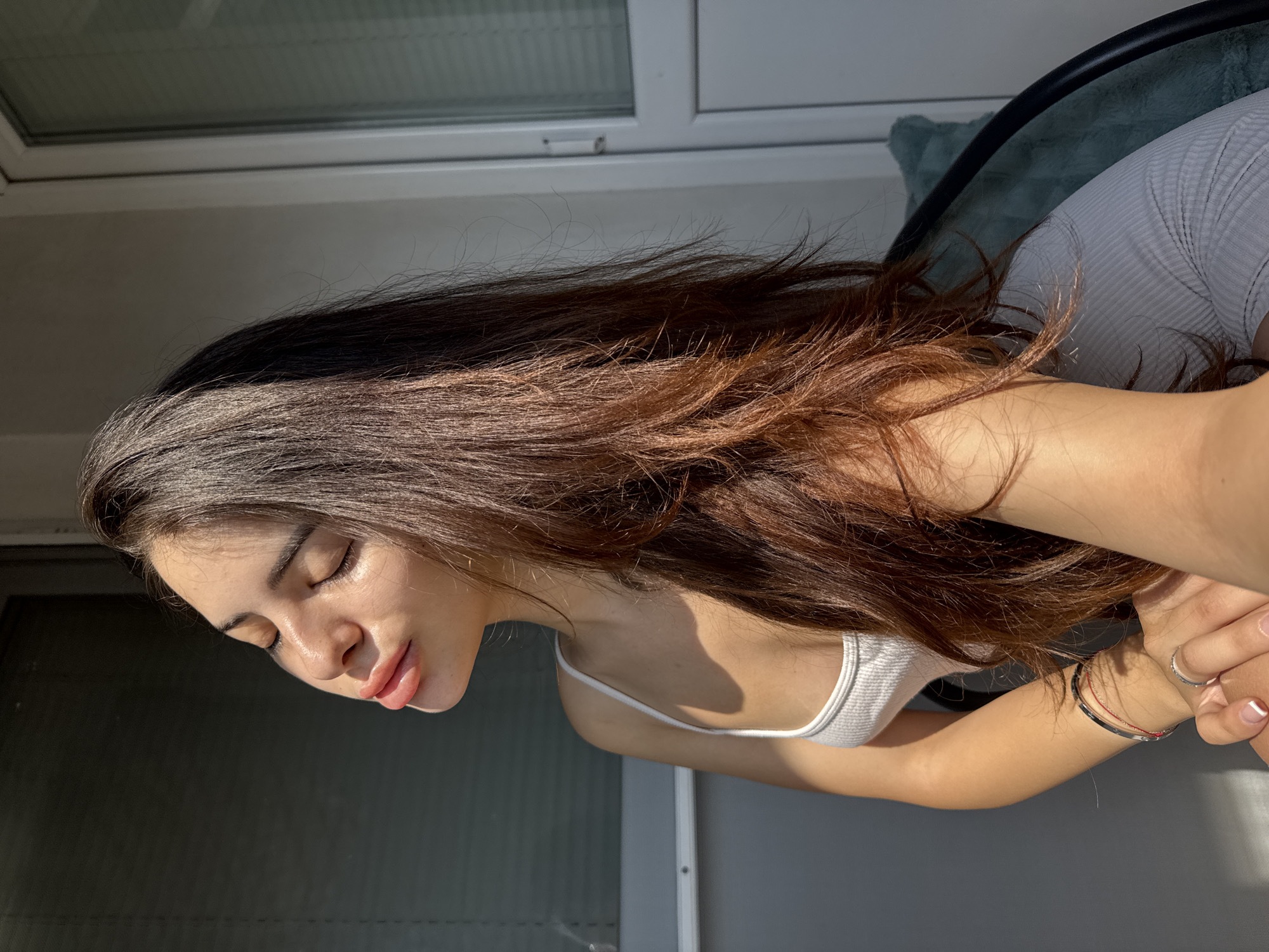 A young adult female is depicted in a sunlit outdoor setting, likely on a balcony or terrace, in Plovdiv, Bulgaria. She has long, dark brown hair with lighter, reddish-brown highlights cascading over her right shoulder and chest. Her eyes are closed, and her head is tilted slightly back and to her left, exposing her face to direct sunlight. Her lips are full and slightly parted. She is wearing a white ribbed spaghetti-strap tank top and a light grey ribbed skirt or shorts. On her visible right wrist, she wears a thin red string bracelet, a silver band bracelet, and another thin dark string bracelet. A silver ring is on her left ring finger, and her fingernails have a French manicure. Her right hand is resting on her left thigh. The background on the left consists of a grey building wall with horizontal blinds. On the right, a white-framed glass door or large window is visible, also with internal horizontal blinds. A black-framed chair with a teal/turquoise fuzzy cushion is partially visible on the far right. Strong sunlight illuminates the scene, casting distinct shadows.