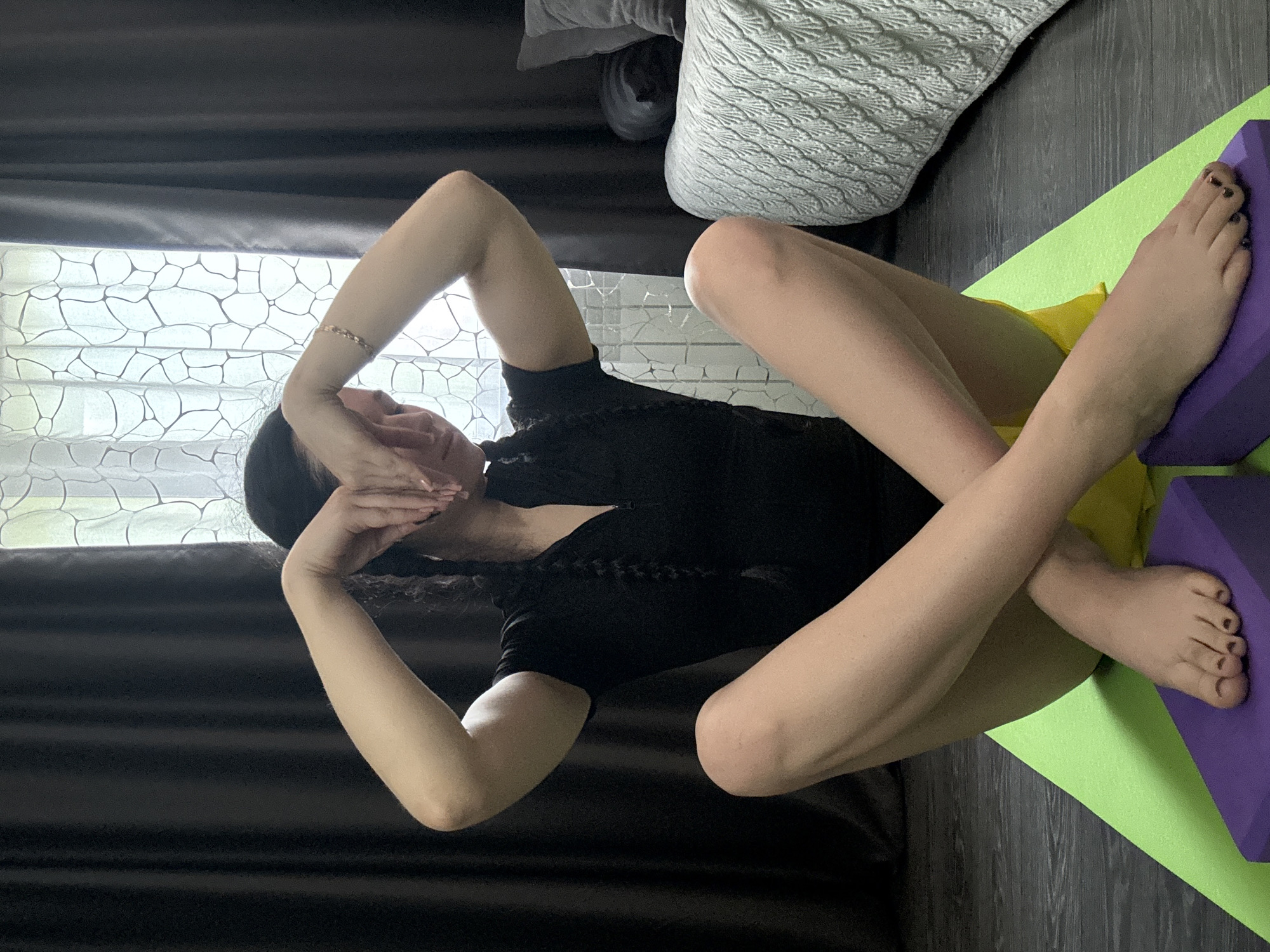 A person with dark hair in braids is sitting on a yoga mat in a lotus position. They are wearing a black zippered shirt and yellow shorts. Their hands are clasped in front of their face. The person's feet are resting on yoga blocks. In the background, there are dark curtains and a window with a patterned sheer curtain. A couch with a textured blanket is visible on the right side of the image. The floor appears to be wooden. This scene is set in a residential interior, likely a living room or bedroom, in Novosibirsk, Russia.