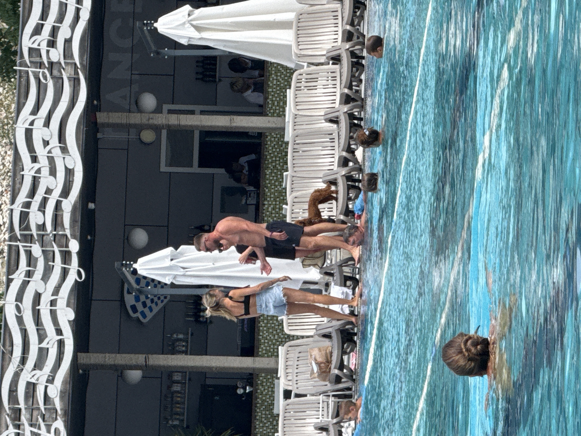 The image depicts an outdoor swimming pool area, possibly a resort or club, with people swimming and lounging. In the foreground, the bright blue water of the pool is visible with several individuals partially submerged. Further back, a woman with blonde hair pulled into a bun swims in the pool. Behind the pool, there is a row of white lounge chairs, some occupied by people. A small dog is standing on one of the chairs. Beyond the lounge chairs is a building with a bar or restaurant area. The building features a decorative element of musical notes above the facade. The setting appears to be a sunny day. The location is specified as Массандровский поселковый совет, Ukraine.