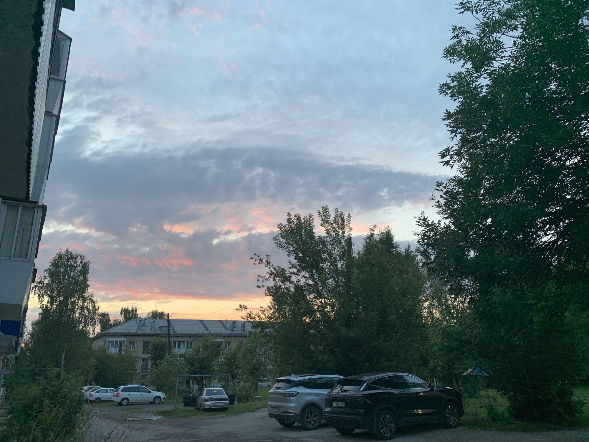 The image depicts a street scene at dusk in the city of Asha, Russia. A multi-story apartment building is visible on the left, with balconies and windows. Several cars are parked in a lot in front of the building, including a silver SUV, a white hatchback, and a black SUV. Tall trees with dense foliage surround the parking area and extend into the background, partially obscuring other buildings and a play structure. The sky is filled with scattered clouds, tinged with pink and orange from the setting sun, with the overall color palette dominated by blues, grays, and greens. The ground appears to be a mixture of gravel and pavement. No people are visible in the image.