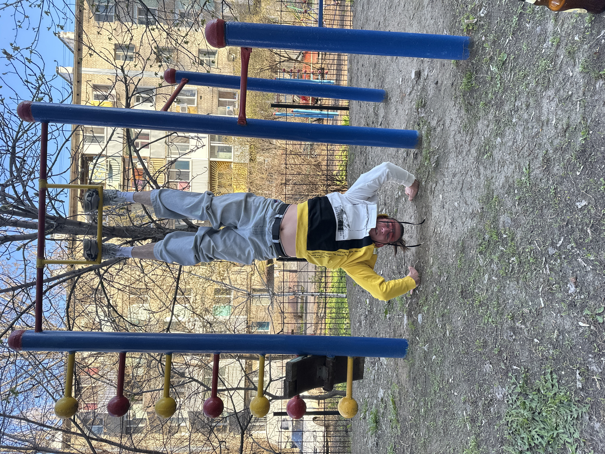 An outdoor, eye-level shot captures a young adult male performing an athletic maneuver on parallel bars in what appears to be a residential playground or exercise area. The subject is positioned horizontally, inverted, with his feet resting on the upper blue bar and his hands gripping the lower yellow bar, demonstrating significant core strength and balance.
He is wearing a yellow and white long-sleeved top with black elements and various printed text, grey baggy trousers, white socks, and white sneakers. His dark hair is styled in thin braids or dreadlocks, and he wears glasses. Visible text on his top includes phrases such as "GOOD VIBES," "LOOKING FOR HOPE," "REPAIR NEW," "LOVE IS ME," and "ABILITY."
The exercise equipment consists of robust blue parallel bars and a lower yellow bar system that includes red and yellow spherical elements. In the background, a multi-story apartment building, constructed from light-colored bricks, rises under a clear sky. The building is partially obscured by the bare branches of trees, suggesting early spring or late autumn. A metal fence runs behind the bars. The ground is a mix of bare earth and sparse patches of green weeds and grass. The time of day appears to be daytime, with bright, natural light. The setting is consistent with a residential area in Saratov, Russia. A brown bottle is faintly visible on the ground near the subject's head. The person's facial expression conveys concentration and effort.