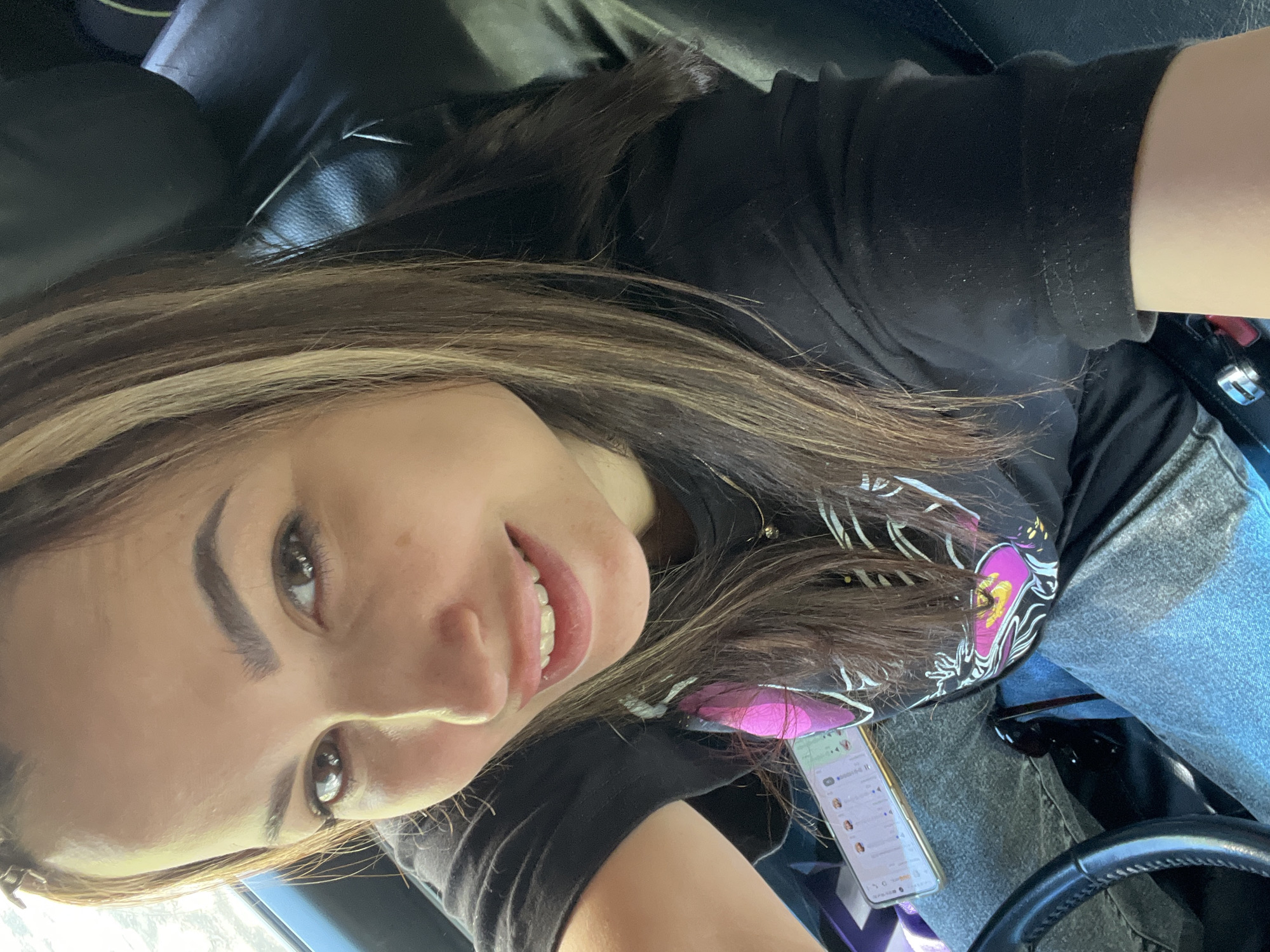 The image shows a young woman in the driver's seat of a car. She is smiling at the camera. She has brown hair with blonde highlights and is wearing a black t-shirt with a graphic design. Her phone is resting on her lap, displaying a messaging app. The background shows the car's interior, including the steering wheel and leather seats. The image appears to be taken indoors. The location is Unknown, Unknown.