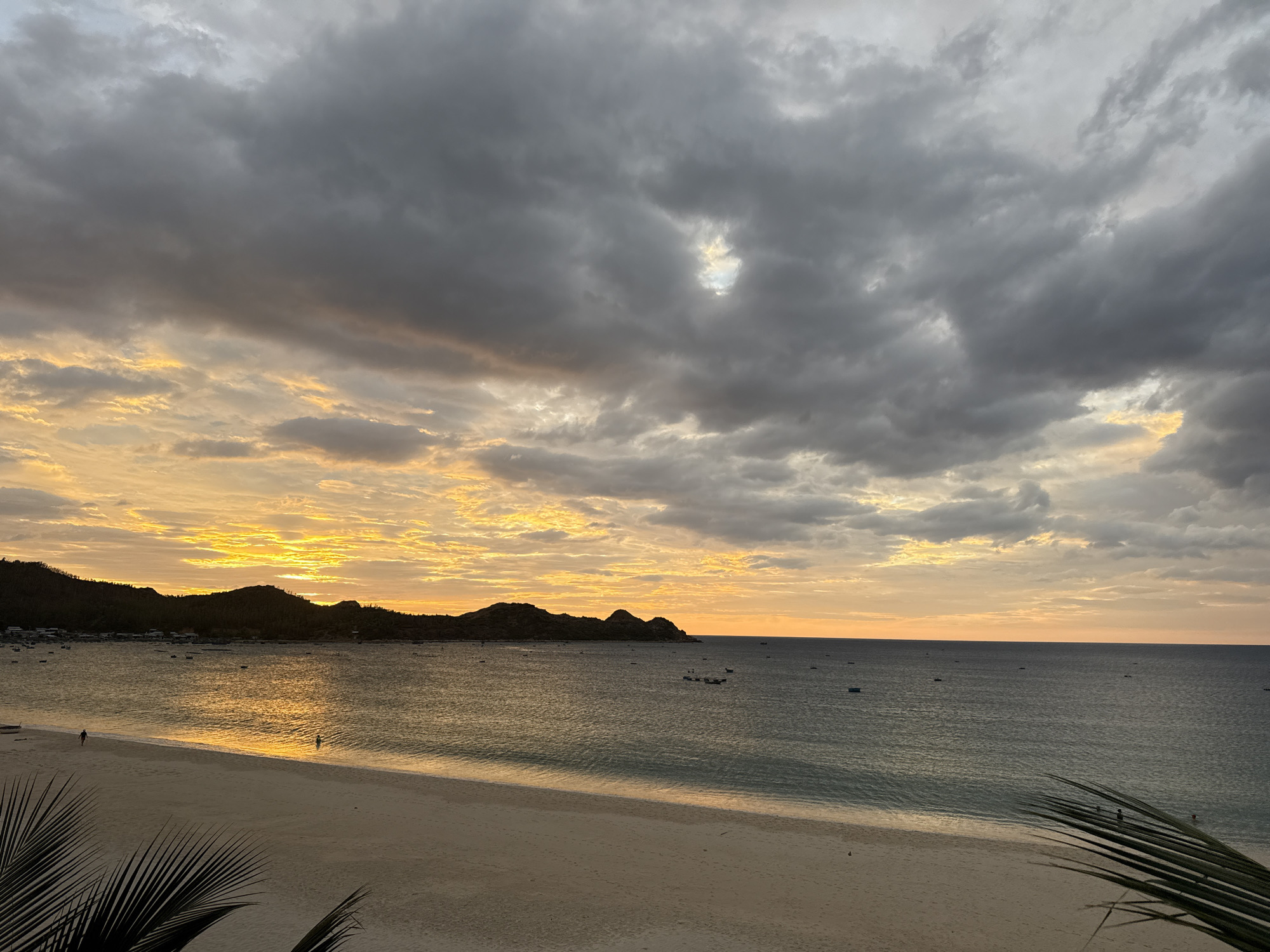 This is a wide-angle, eye-level, horizontal shot of a beach at sunset in Song Cau, Vietnam. The sky is filled with dramatic, dark gray clouds that are illuminated by the setting sun, creating a vibrant orange and yellow glow. A strip of golden light reflects on the water. The sea is calm with small waves rolling onto the shore. The beach is sandy and mostly empty, with two people visible in the shallow water near the shoreline. In the distance, a silhouetted line of hills forms the coastline, dotted with small structures and boats. In the foreground, the fronds of a palm tree are visible on the right side of the frame.