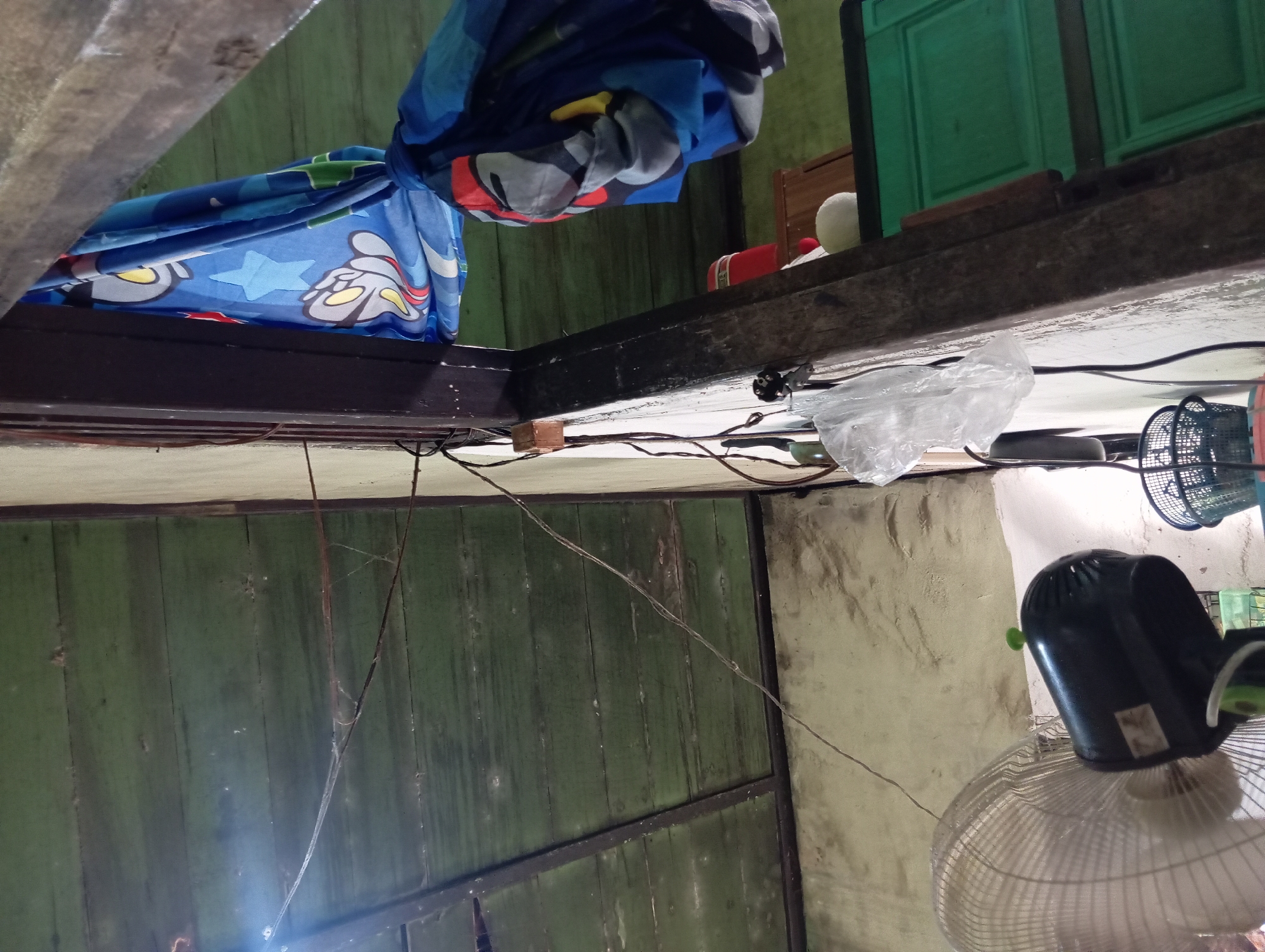 The image depicts an indoor scene, likely a room in Rejosari, Indonesia. The view is angled upwards, focusing on the ceiling and a vertical wooden beam. The ceiling is constructed of aged, horizontally placed wooden planks painted green, showing signs of wear and some cobwebs.
Attached to the wooden beam are several electrical wires, some of which are thin and taut, others thicker and draped. A small, rectangular wooden block is affixed to the beam, likely serving as a conduit or support for the wiring.
Hanging from the beam is a piece of blue fabric with a cartoon pattern, featuring characters like Mickey Mouse and stars. The fabric is gathered and tied, suggesting it might be a curtain or part of a textile covering.
Below the beam and wires, a white wall with a textured surface is visible. To the left, a partially visible electric fan with a white grille and a black motor is positioned. Its blades are not in motion. Next to the fan, a black wire is connected to a power outlet. A small, black basket with a woven pattern is also present.
Further to the right, a tall, dark green cabinet with patterned doors is visible. On top of the cabinet, a red cylindrical object and a white spherical object are present. The overall impression is of a functional, somewhat cluttered domestic interior.