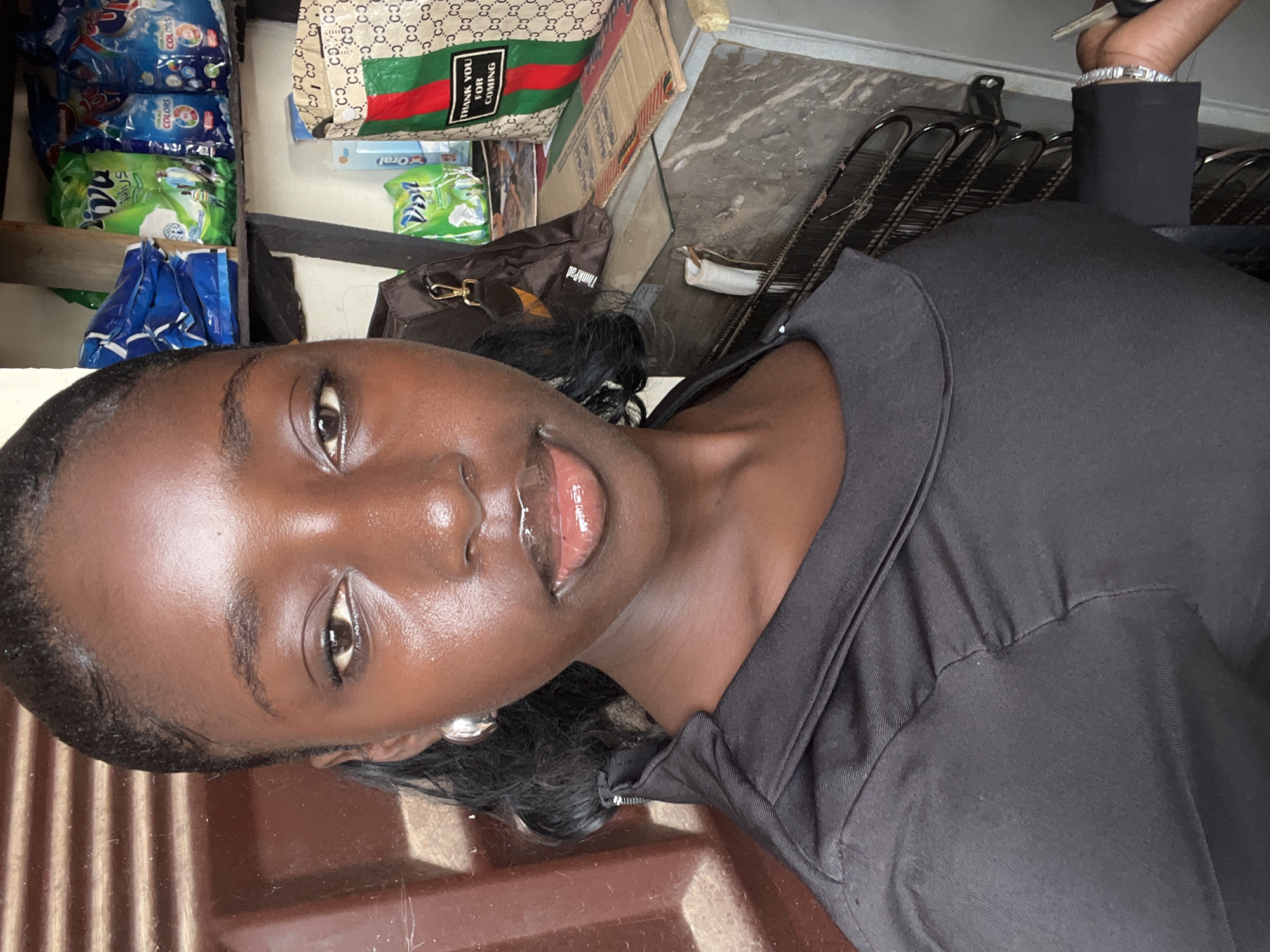 A dark-skinned young woman is seen from a close, slightly elevated angle, likely a selfie. She wears a dark-colored top and her hair is styled with some strands near her face. Behind her, a variety of objects are visible, including packages of detergent or cleaning supplies, a designer tote bag with "THANK YOU FOR COMING" printed on it, a brown "ThinkPad" bag, and metal racks. The scene suggests an informal setting, possibly a home or small business. There is no information in the image to indicate the location is Abuja, Nigeria.