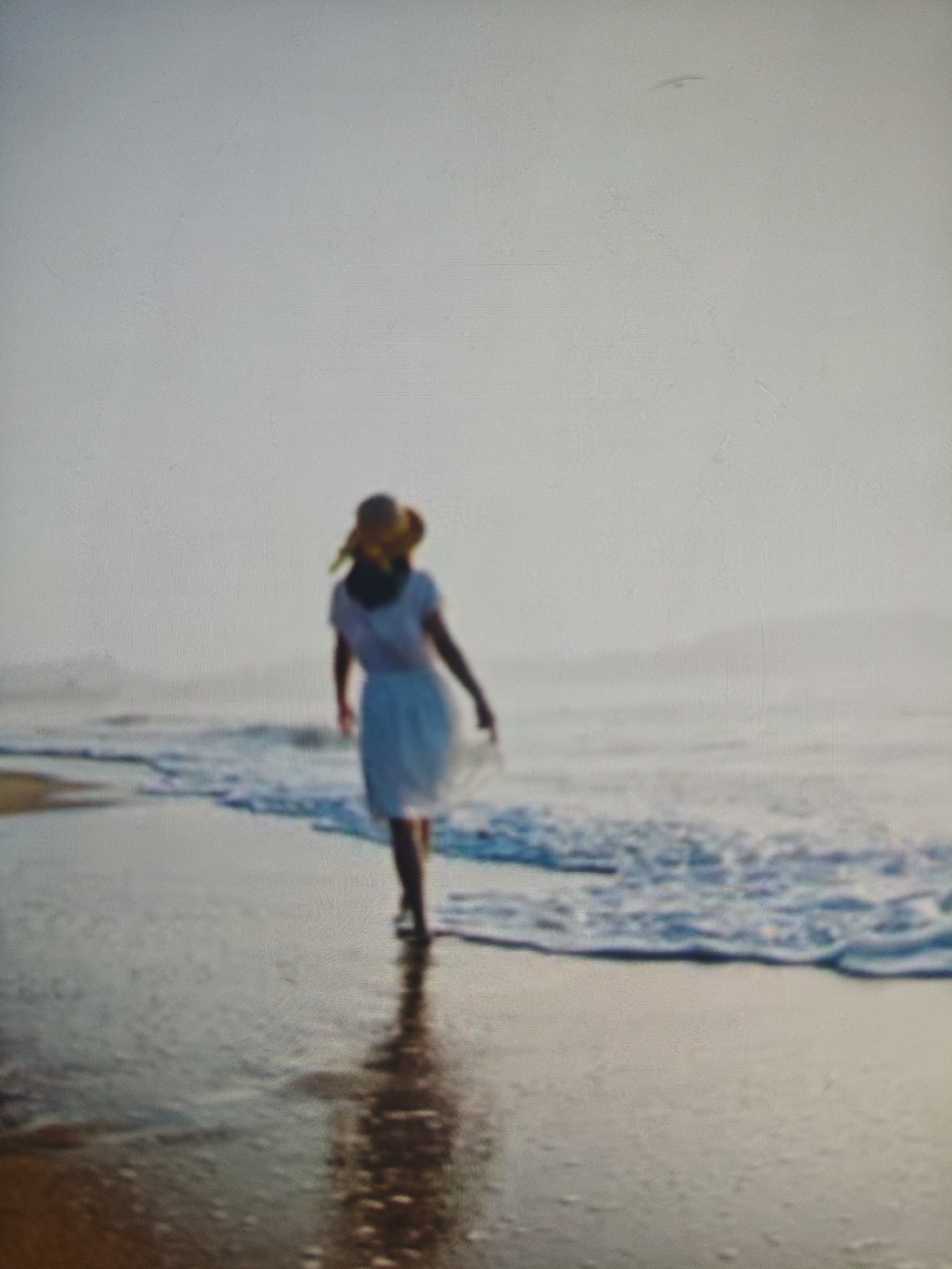 A full-shot, blurred photograph shows a woman walking away from the camera on a wet, sandy beach. The woman, wearing a white dress and a straw hat, is walking towards the water, with waves lapping at her feet. The sky is overcast, and the sea is calm. There is no visible indication of the location being Krasnaya Polyana, Russia.