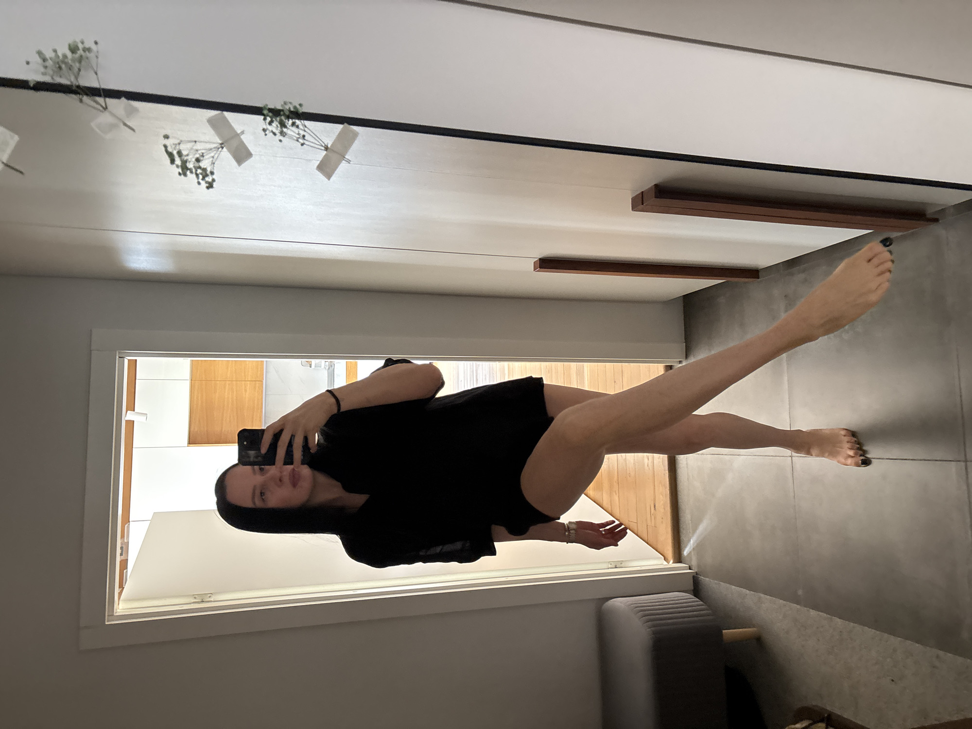 A young woman is captured in a mirror selfie taken indoors in Chaikovsky, Russia. She has long, straight dark hair and a neutral expression, looking directly at her smartphone, which she holds in her left hand. She is wearing a loose-fitting black t-shirt or oversized top, and her bare legs are visible. Her left leg is straight, while her right leg is bent and raised, resting against a surface within the reflected scene.
The reflection in the mirror reveals a modern interior space. Along what appears to be the top edge of the reflected room, several small sprigs of light-colored baby's breath flowers are taped to the wall with white tape. Below these flowers, two horizontal dark wooden planks or decorative beams are mounted on the reflected wall. The floor visible in the reflection, and also in the foreground of the actual room, is composed of large, light grey tiles. Part of a grey upholstered bench or sofa is visible in the lower right foreground, outside the mirror's reflection. The lighting is bright, suggesting daytime. The woman's toenails are painted dark. There is no visible text in the image.