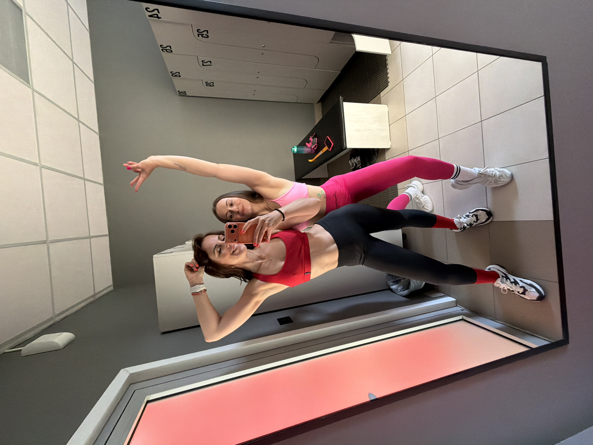 Two women are taking a mirror selfie in a locker room. The woman on the left is wearing a red sports bra and black leggings with white sneakers and red socks. She is smiling and holding her phone up to take the photo. The woman on the right is wearing a pink sports bra and pink leggings with white sneakers and white socks. She is also smiling and has her arm raised in the air. In the background, there are white lockers and a black bench with a water bottle and a phone on it. The floor is tiled and the walls are gray. The scene appears to be in a gym or fitness center.