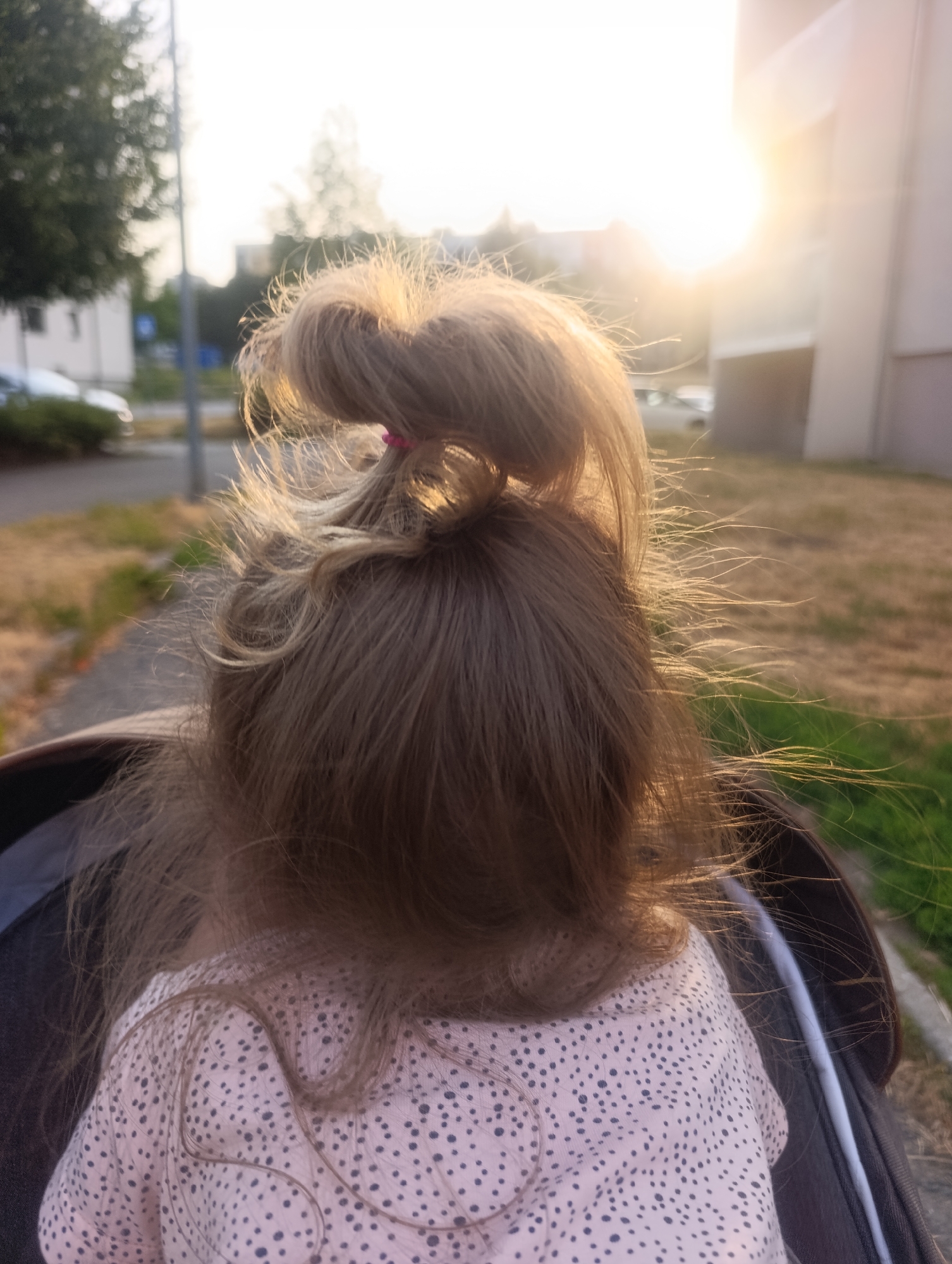 The image shows the back of a young child's head, with her hair pulled up in a messy bun. The child is wearing a pink shirt with black polka dots. She is sitting in a stroller, and the background is blurred, showing a street scene with a building and trees. The sun is shining brightly, creating a lens flare in the upper right corner of the image. There is no clear indication of the specific location within Olomouc, Czechia, or any specific action or interaction occurring beyond the child being in the stroller.