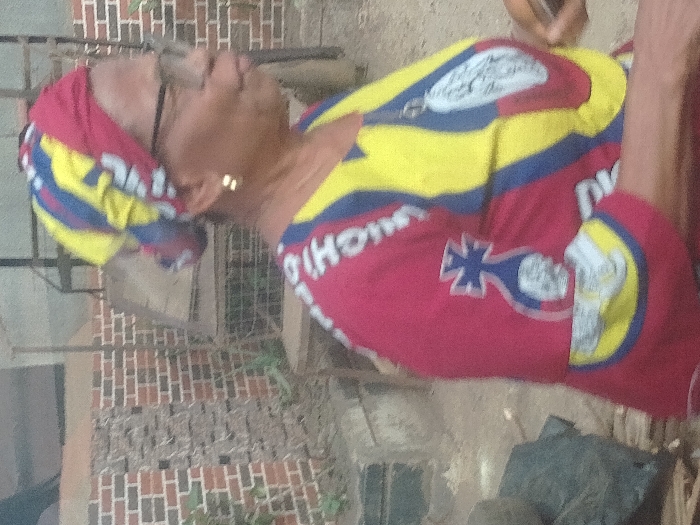 The image displays an outdoor scene in Orie Uratta, Nigeria. An adult female, visible from the upper torso in profile, faces toward the right. She wears a multi-colored headscarf featuring patterns in red, yellow, and blue. Her attire includes a matching multi-colored top with dominant red and yellow sections, blue stripes, and white graphical elements, including a distinct blue cross design over a white shape on the left shoulder area and partial white text "GHOMA". She wears clear-framed eyeglasses and small gold-toned stud earrings. Her mouth is slightly open. Her right hand, partially visible in the lower right, holds a dark rectangular object, consistent with a smartphone. The background features a red brick wall on the left, an unpaved ground surface, and various metallic structures, some appearing as frames or furniture components. Sparse green vegetation is visible near the brick wall.