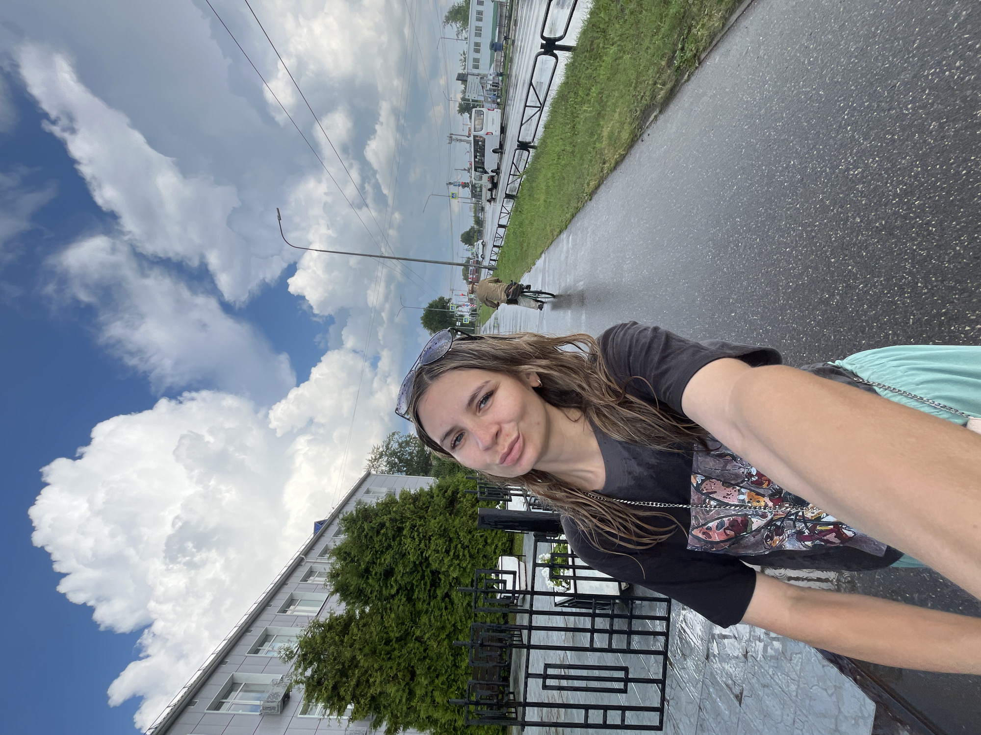 A young woman is taking a selfie on a wet sidewalk in Chaikovsky, Russia. She is wearing a black t-shirt with a colorful graphic and light blue shorts. She has long, wavy brown hair and is wearing sunglasses on her head. Behind her, there is a black metal fence and manicured evergreen trees. In the background, a road is visible with traffic, including a bus and a person riding a bicycle. The sky is cloudy with large, white cumulus clouds. The sidewalk and road appear wet, suggesting recent rainfall. The buildings in the background are typical of Soviet-era architecture.