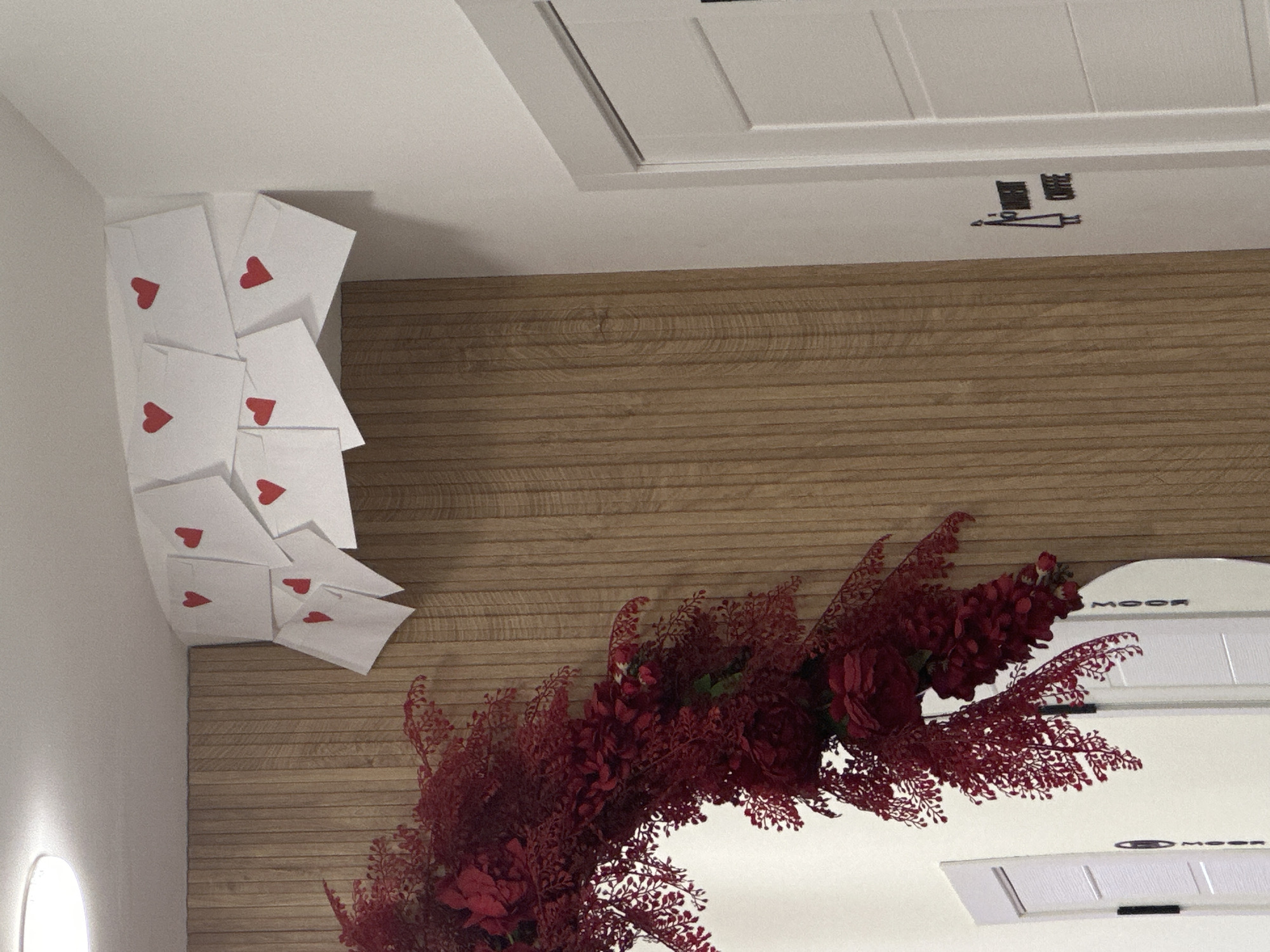 A slightly rotated, upward-angle shot reveals a decoratively adorned interior space, presumably a business in Yerevan, Armenia. The ceiling, painted white, features a recessed rectangular panel with interior molding. Affixed to a lower wooden-paneled wall are scattered white envelopes, each marked with a red heart. A lush arrangement of dark red flowers and foliage is positioned toward the bottom right. Part of a white appliance with "MOOR" printed is seen behind the floral arrangement.