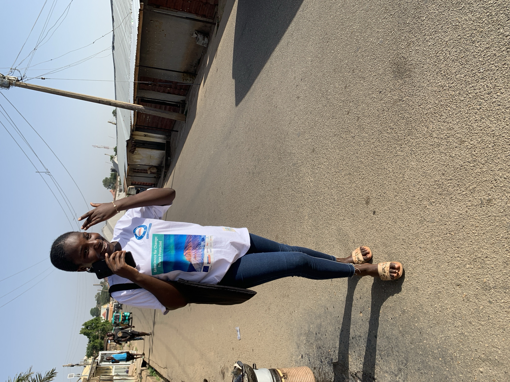A woman stands on a paved street in Jos, Nigeria, talking on a mobile phone. She wears a white T-shirt with a "Geoscience for Change: Future Protected" logo, blue jeans, and sandals. Overhead power lines are visible, along with buildings. The lighting suggests a sunny day.