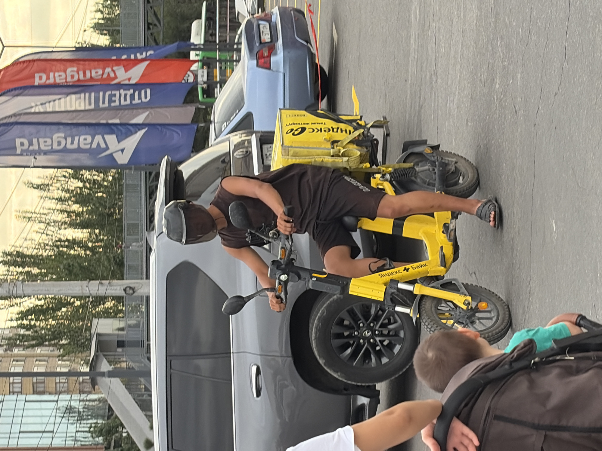 In an urban outdoor setting, a person wearing a dark sleeveless shirt, dark shorts, and dark sandals rides a yellow electric scooter. The scooter has a large yellow insulated delivery box mounted on the rear. The person wears a dark helmet that obscures their face. Behind the scooter, a dark-colored SUV is parked. To the right of the SUV is a blue sedan. In the background, there are several tall vertical banners, one of which displays the text "Vanguard". Further in the background, a pedestrian overpass and trees are visible. The ground is paved with asphalt. A partial view of a stroller and a child's head are visible in the foreground on the left side of the image.