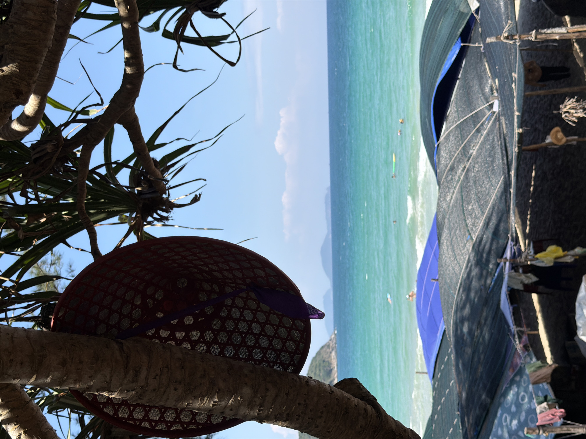The image shows a coastal scene in Cam Lam, Vietnam. The view includes a tree branch with a woven hat resting on it in the foreground. In the background, turquoise water meets a light blue sky with a few scattered clouds. There appear to be some people enjoying activities in the water. To the right, a shaded structure or shelter is visible. The image is rotated counter-clockwise.