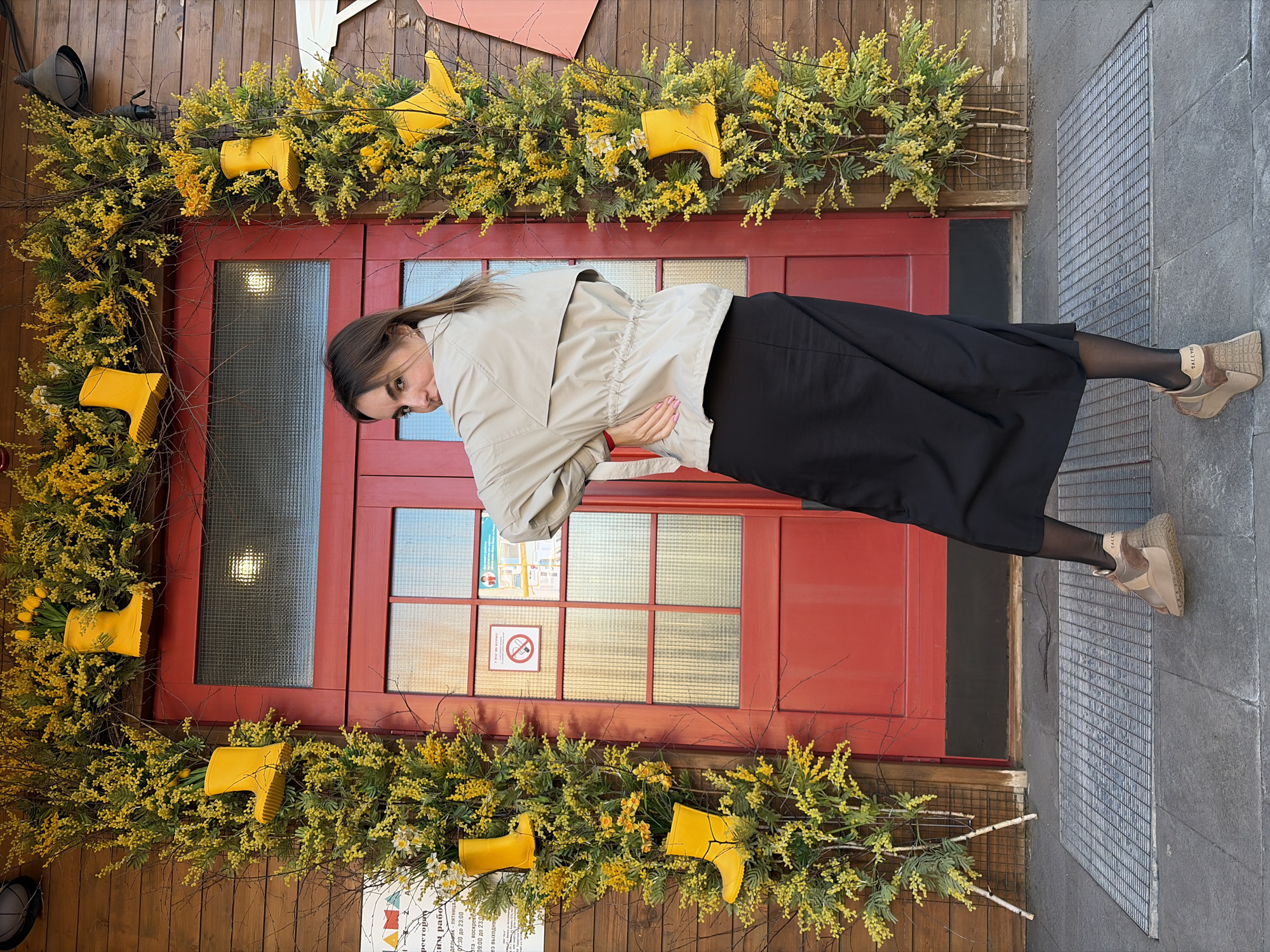 A woman in a black skirt and beige jacket stands in front of a red door decorated with yellow flowers and yellow rubber boots, posing for a photo. The door is part of a wooden building, with an informational sign visible at the bottom. A metal grate lies on the ground to the right of the door. The text on the sign mentions a restaurant and work hours. The location is likely Izhevsk, Russia, based on the Cyrillic text on the sign.