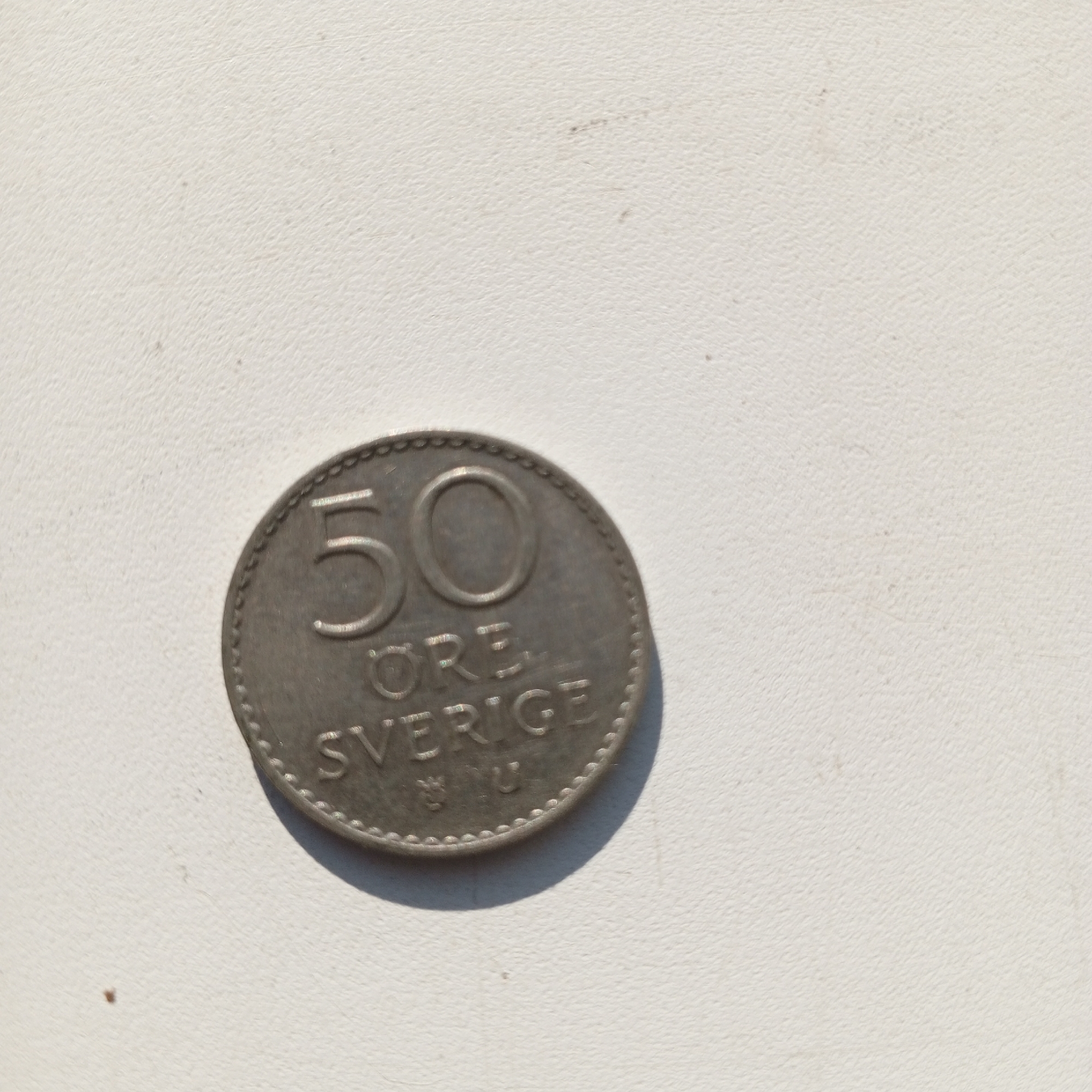 The image shows a Swedish 50 öre coin placed on a textured white surface. The coin is silver-colored and shows the number "50" prominently on its obverse side, along with the text "ÖRE" and "SVERIGE." The surface appears to be a piece of textured paper or fabric. There is no visible indication of the city of Sayanogorsk, Russia, or any other specific location. The lighting is overhead, casting a slight shadow to the right of the coin. The scene is static and there are no people or actions depicted.