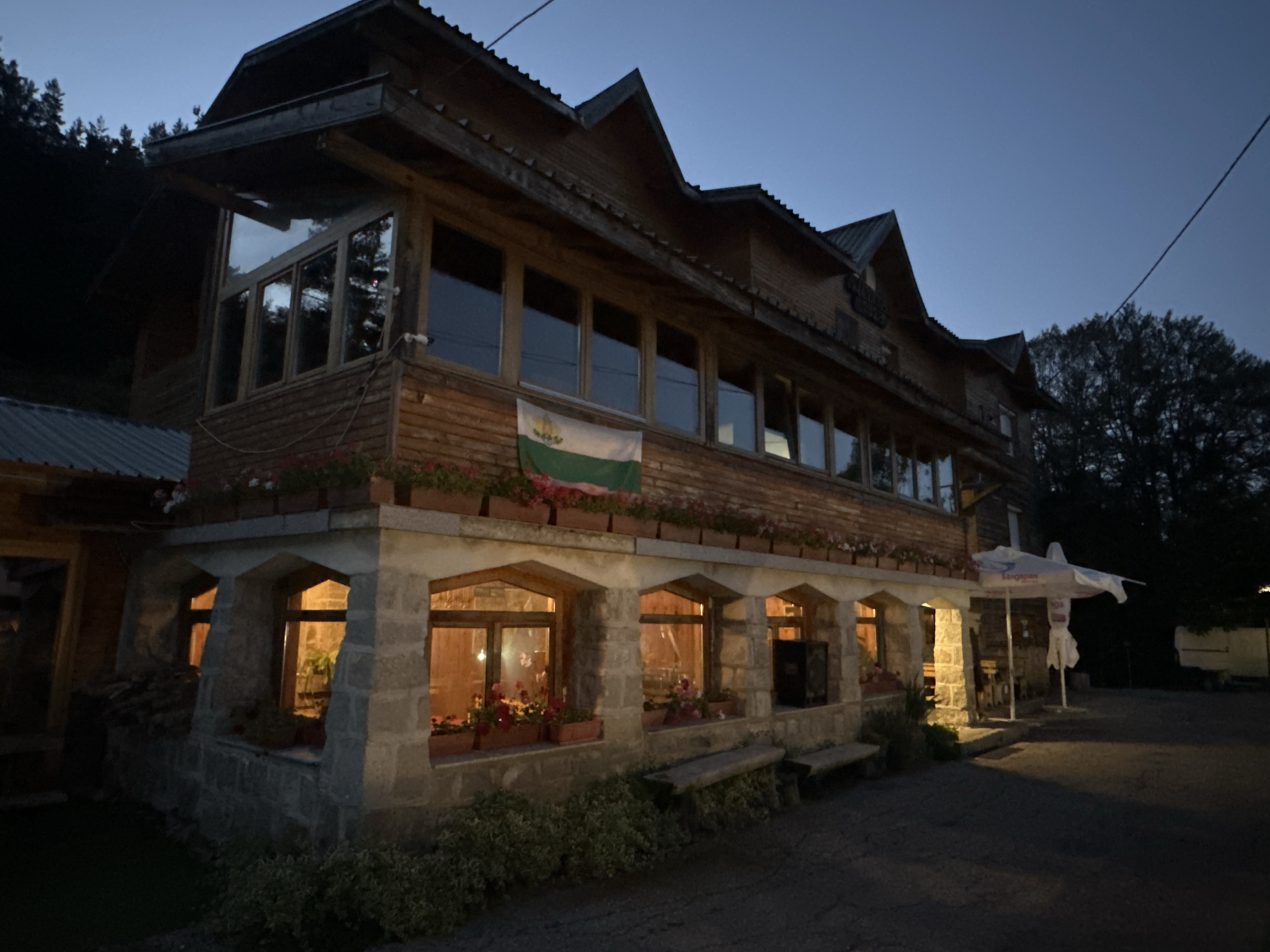 The image depicts a two-story wooden building with stone accents, possibly a guesthouse or restaurant, located in Bogoslov, Bulgaria. It is dusk, and the sky is a deep blue. The building features multiple windows on both levels, some of which are illuminated from within, casting a warm glow. Flower boxes filled with red and pink flowers adorn the exterior, adding a touch of color. A Bulgarian flag is prominently displayed on the second floor. The foreground shows an outdoor seating area with wooden benches, and beyond that, a dark asphalt surface. Trees and foliage surround the building, and a power line is visible stretching across the sky. In the background, a white umbrella and what appears to be a trailer are partially visible. The overall scene conveys a rustic and inviting atmosphere.