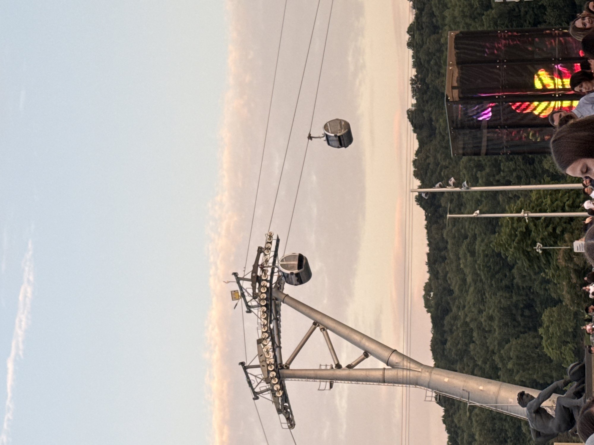 In Moscow, Russia, this image captures an outdoor scene with a cable car system as a prominent feature. Two cable cars are visible, one attached to a large support tower and another suspended on a cable. The support tower is metallic and industrial in appearance, with visible machinery at the top. In the background, a densely wooded hillside stretches across the frame, suggesting a natural area within or near the city. In the foreground, a crowd of people is gathered, indicating an event or public gathering. A statue, likely of a male figure, is also visible in the lower left portion of the image. To the right, a dark, cubic structure with colorful lights emitting from within suggests a stage or a light installation, further implying a public event. The sky above is a pale blue with soft, wispy clouds tinged with pink and orange, indicative of a sunset or sunrise.
