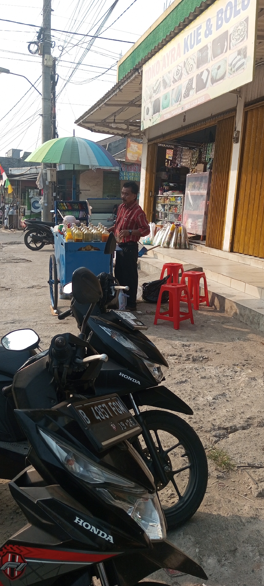 In Setu, Indonesia, a street vendor stands by a blue cart filled with bottles of yellow liquid, likely beverages, under a colorful umbrella. He is wearing a red and white striped shirt and black pants and appears to be serving a customer or arranging his wares. Two black Honda motorcycles are parked in the foreground, one with a license plate reading "B 4957 FGM 19-26". In the background, a shop with a yellow awning displays a sign that reads "TOKO AYRA KUE & BOLI", indicating it sells cakes and bread. The scene depicts a typical street-side commercial activity with food and beverage vendors and nearby shops.