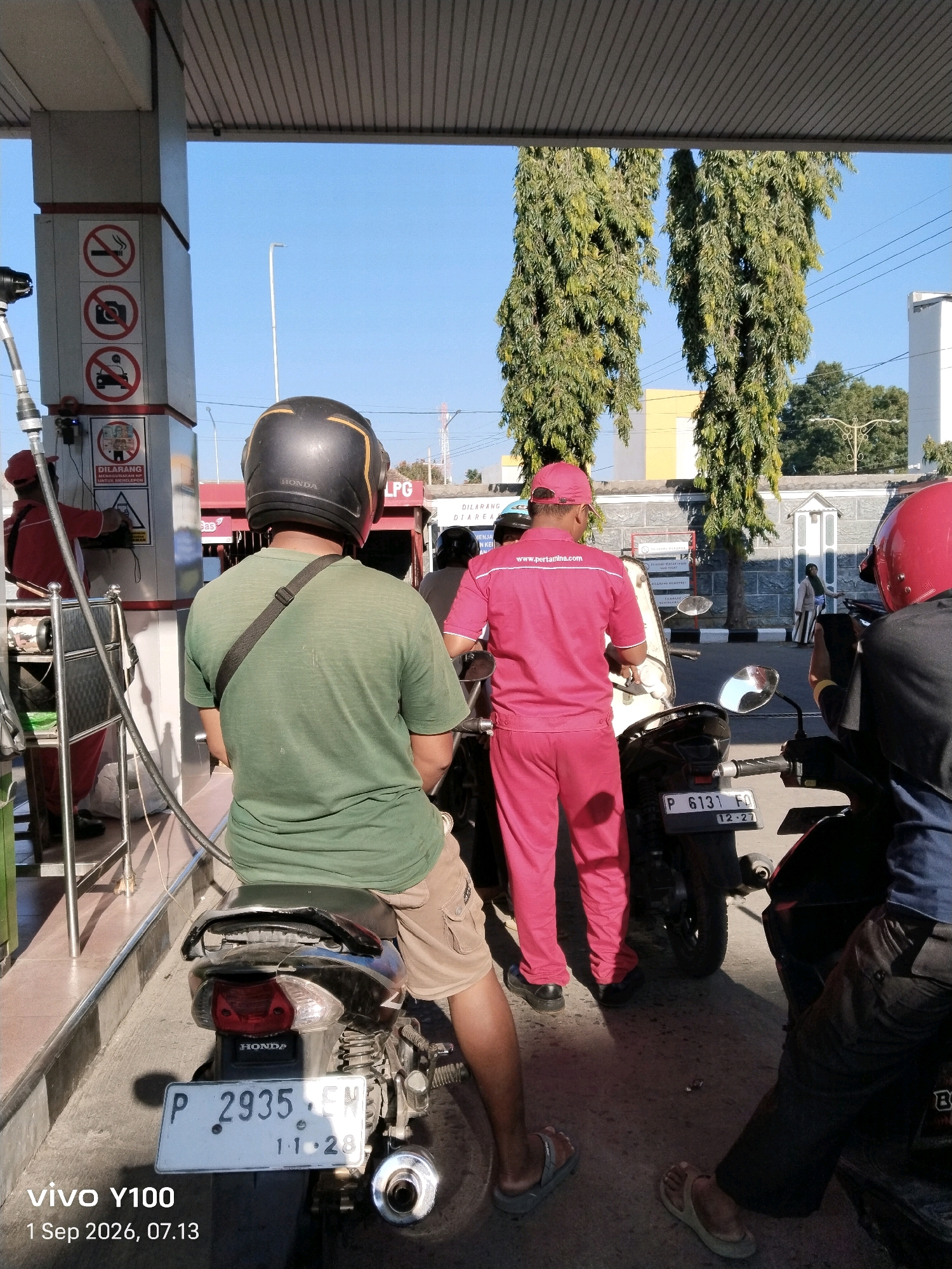 In Situbondo, Indonesia, at what appears to be a gas station, several individuals are present with their motorcycles. One person, wearing a green t-shirt and khaki shorts, is seated on a motorcycle, facing away from the camera, with a black helmet on. Another person, in a bright pink uniform with the "www.pertamina.com" logo, stands near a gas pump, facing a motorcycle with its license plate visible as "P 6131 FQ 12-27". A third individual, in a red helmet, is also on a motorcycle, with their back to the camera. The environment includes a gas pump structure with caution signs, tall trees, and buildings in the background under a clear blue sky. The ground is paved asphalt.