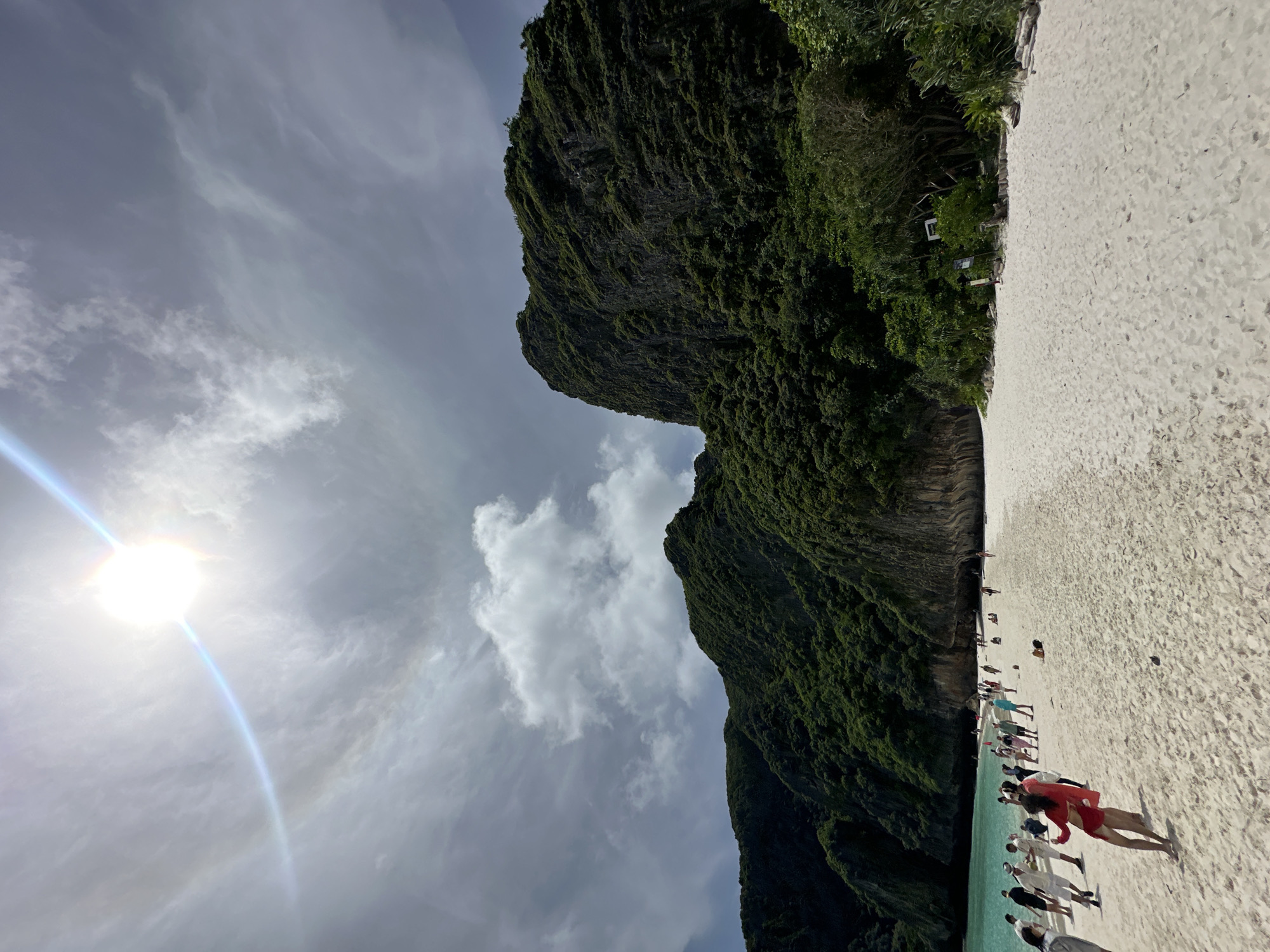 The image depicts a scenic beach with a rocky, lush green cliff face dominating the left side of the frame. The foreground shows a sandy beach with several people scattered across it, some walking near the turquoise water and others further up on the sand. The sky is partly cloudy with the sun visible, creating a lens flare and a faint rainbow arc. The overall scene suggests a tropical beach destination, likely during the daytime. The orientation of the image is rotated 90 degrees clockwise. People are dressed casually, indicating a relaxed atmosphere. The presence of multiple individuals suggests it's a popular tourist spot. No visible text is present in the image. The location context of Ao Nang, Thailand aligns with the visual cues of a tropical beach with limestone cliffs.