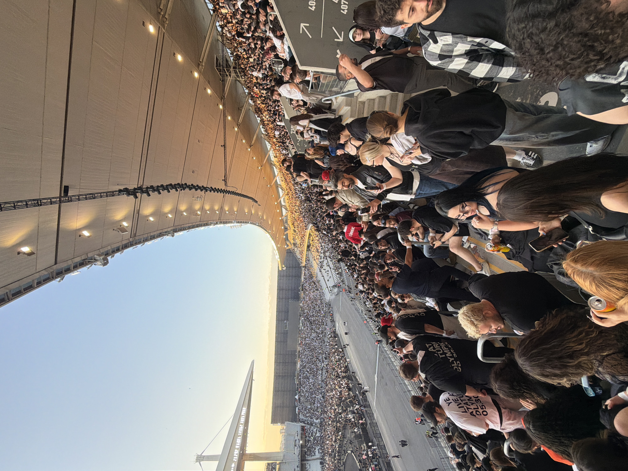 The image shows a large crowd of people at Başakşehir Fatih Terim Stadium, located in Başakşehir, Turkey. The crowd fills the stadium stands, with many people standing close together. There's a large structure visible to the left, with the sky behind it. People in the foreground are wearing casual clothing, and some are holding cans or phones. Signs are visible, including one that says "YE LIVE ATATÜRK OLYMPIC 05.30." There are also signs with the section numbers 405 and 407.