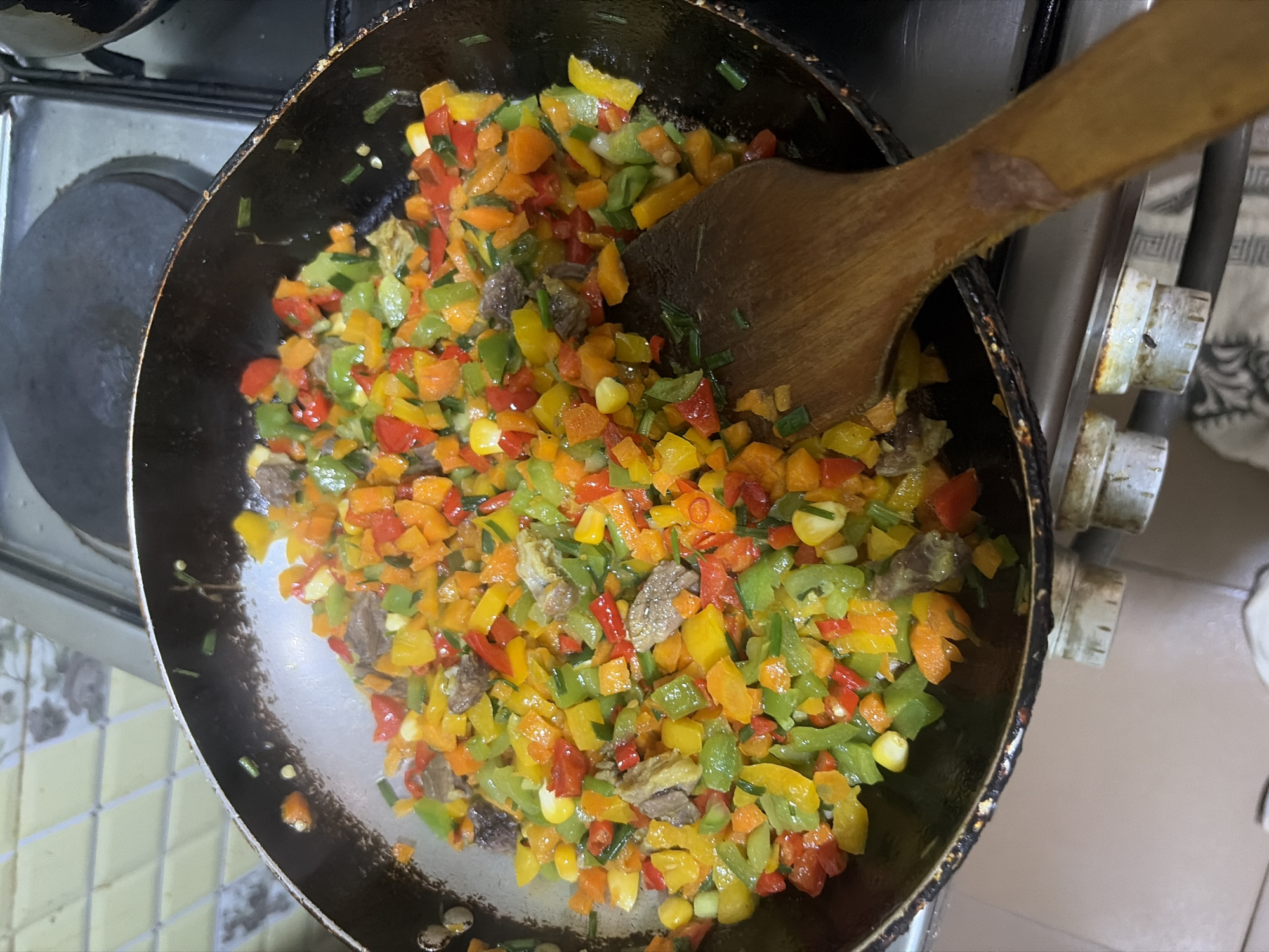 The image captures an overhead view of a black frying pan situated on a stovetop. The pan contains a mixture of diced red, yellow, and green bell peppers, orange diced vegetables, small pieces of cooked brown meat, yellow corn kernels, and chopped green herbs. A wooden spatula is partially submerged in the food, positioned towards the lower right portion of the pan, indicating stirring. The pan's interior and rim show visible signs of use. The stovetop surface includes dark circular elements and a metallic grate on the right. Below the pan, two white cylindrical control knobs with numerical markings are visible. The background features light yellow kitchen tiles with a diamond pattern. No human figures are present. The location is the city of Unknown, Unknown.