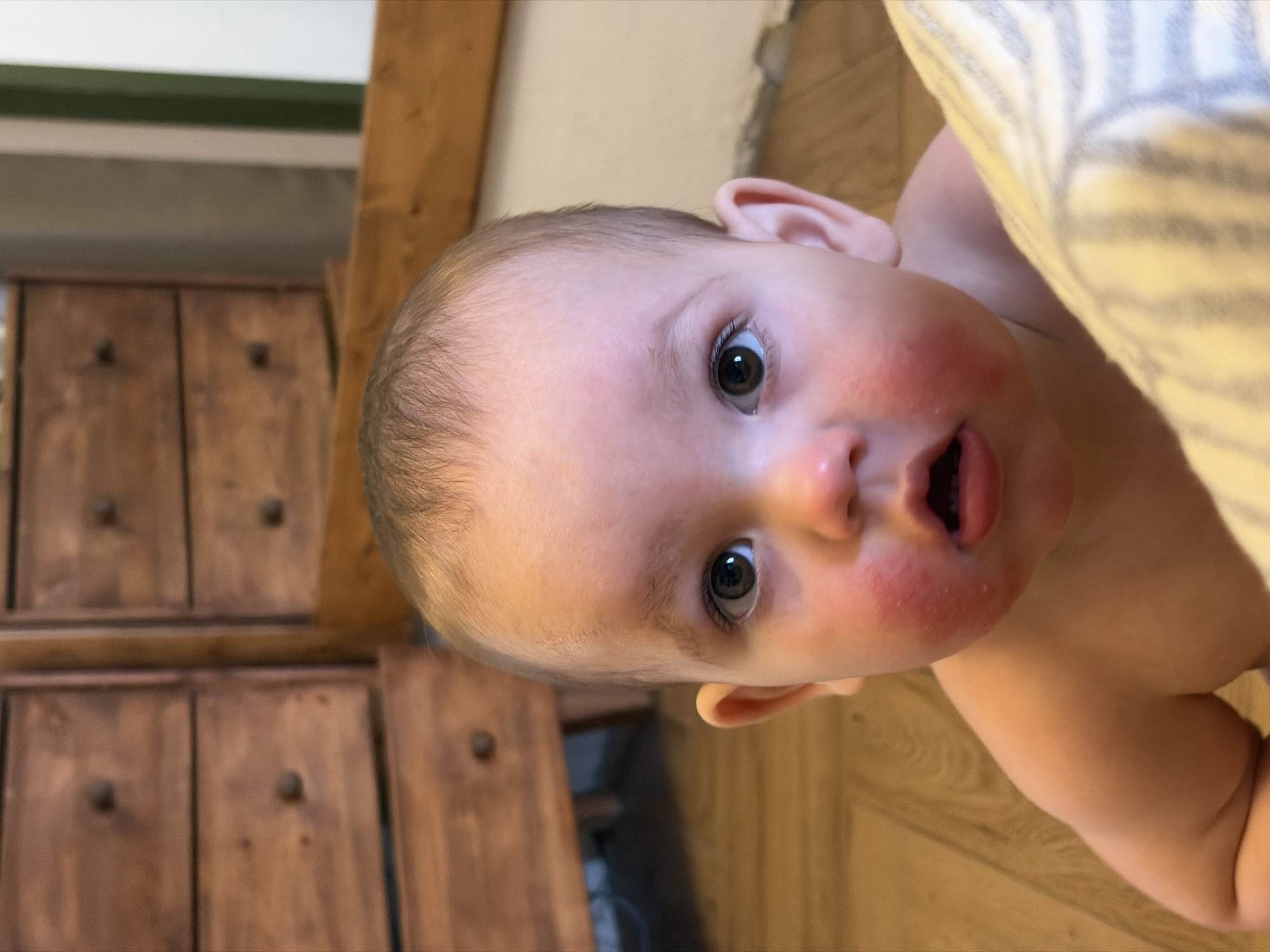 A close-up, eye-level shot depicts a baby with a surprised expression, facing upwards with an open mouth. The baby has rosy cheeks and dark eyes. Part of their body is visible, draped with a striped fabric, possibly a blanket. The background includes a partially visible wooden cabinet with dark knobs and a wooden floor. The specific location in the city of Chaikovsky, Russia, is not discernible from the provided image.