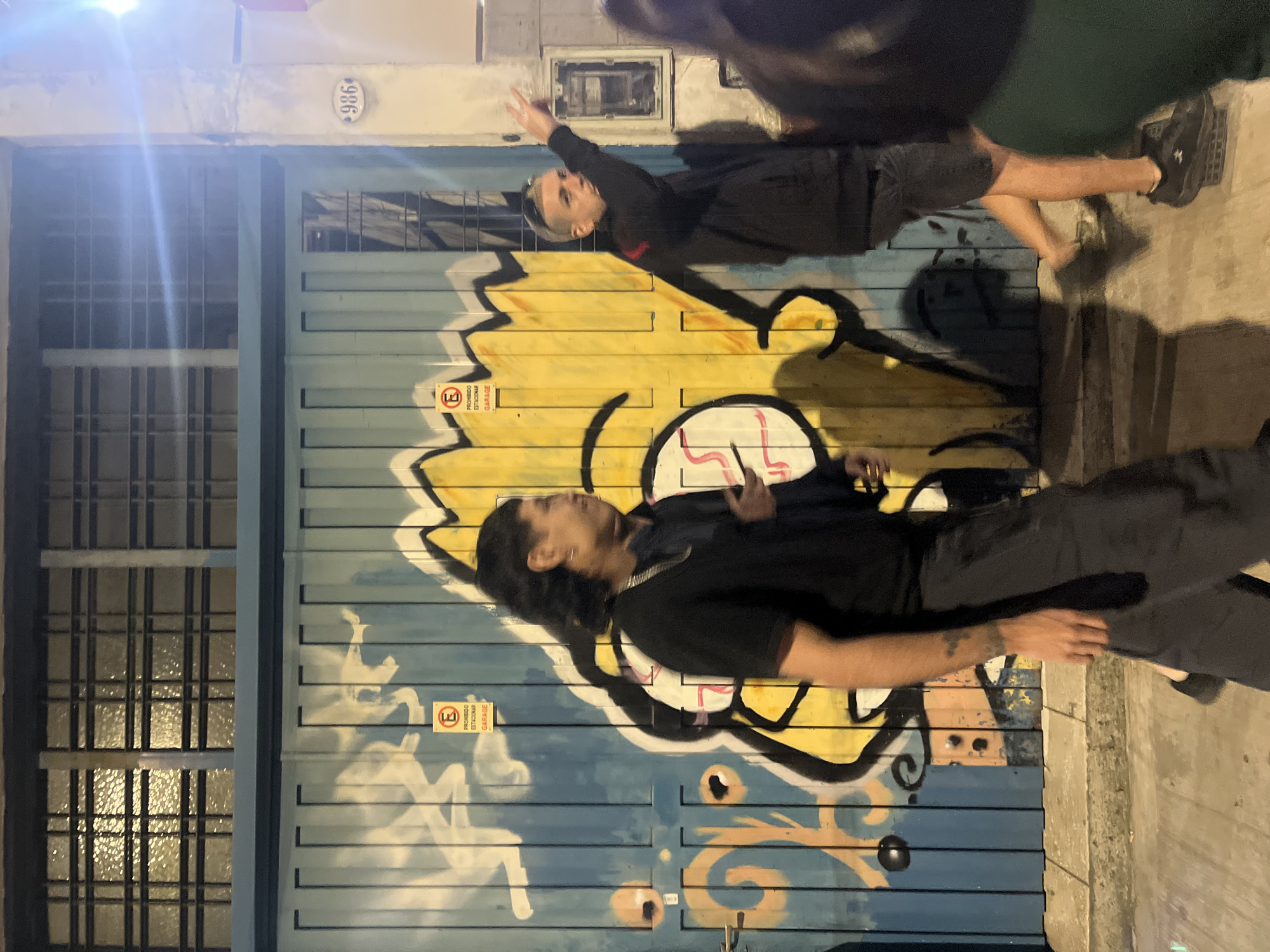 The image shows two people standing in front of a roll-up garage door covered in graffiti. The first person has long, dark hair and is wearing a black shirt and dark pants. The second person has short, light hair and is wearing a black jacket and shorts. Both are gesturing with their hands. The garage door is painted blue and yellow, with black outlines, and features a no parking sign. To the left of the garage door is a window with a metal grate. Above the door is a sign reading "986". The location is likely in Buenos Aires, Argentina.