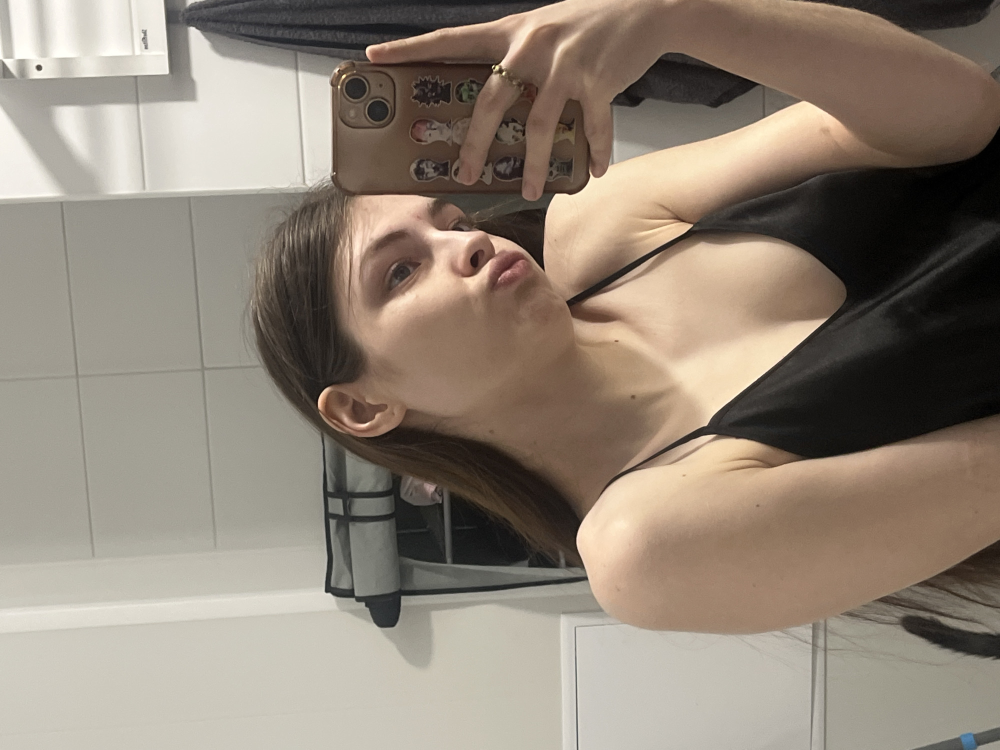 A young woman is taking a selfie in what appears to be a bathroom. She is holding a smartphone with stickers on the back in her right hand. She has long brown hair, and is wearing a black top with thin straps. The background includes white tiled walls, a towel rack and a mirror. There is no indication of the city of Saint Petersburg, Russia, or any information to determine its location.