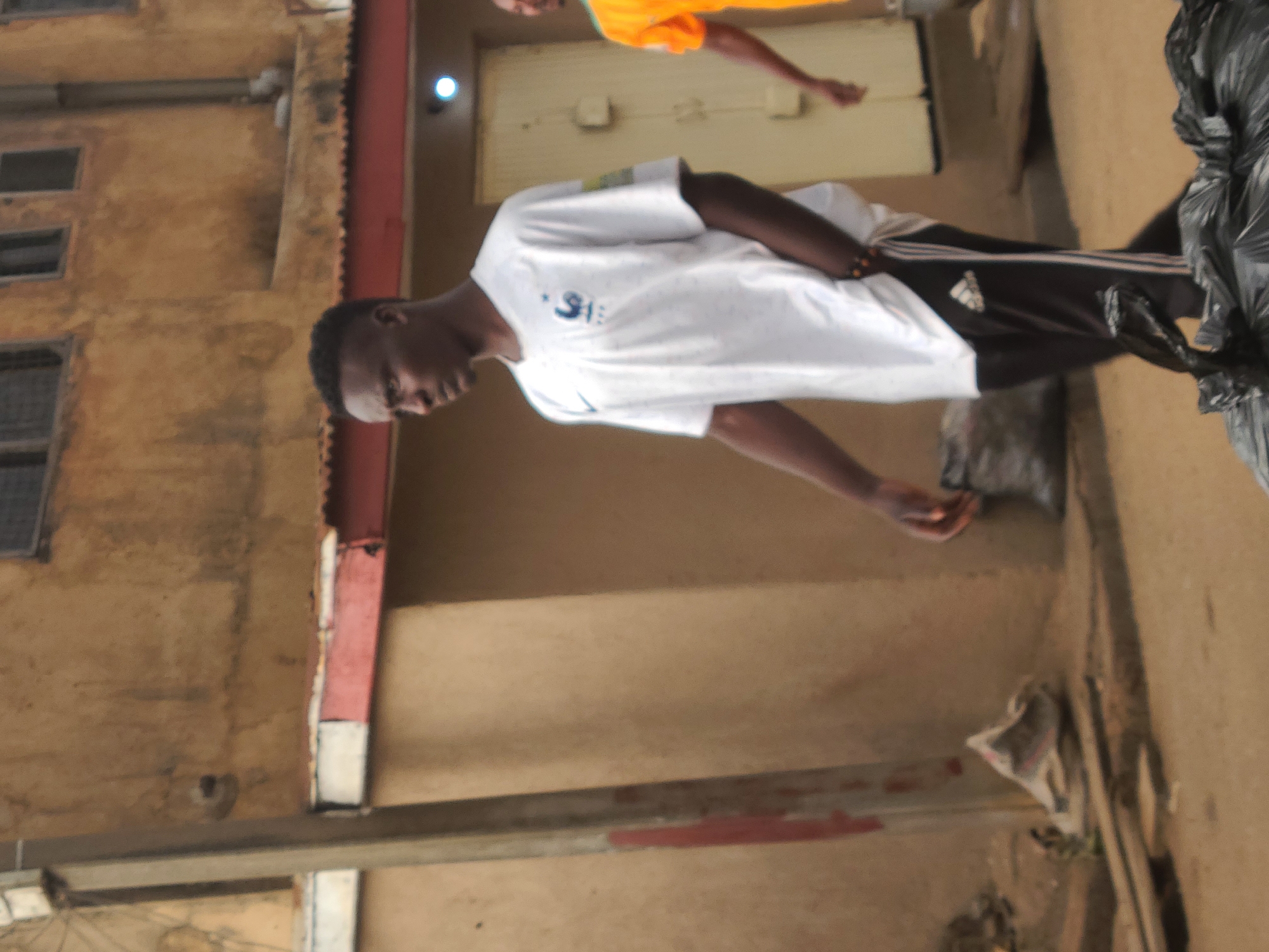 The image captures an urban scene, possibly a street or alleyway, within Jos, Nigeria. A young man, wearing a white t-shirt and black Adidas track pants, stands prominently in the foreground. The background features weathered, multi-story buildings with visible windows and architectural details. Red-painted structural elements, like pillars or beams, are part of the buildings' framework. In the immediate surroundings, there are signs of everyday life and urban activity, including objects wrapped in black plastic, and scattered debris. The overall setting appears to be a bustling, densely populated area of Jos.