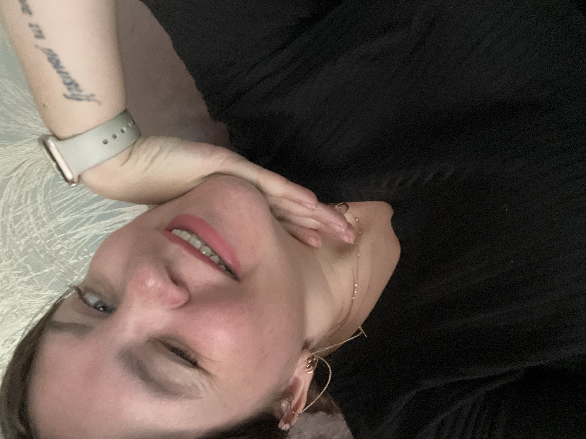 This is a close-up selfie of a woman in Noviy, Russia. She is looking at the camera with a wink and a smile. She is wearing a black shirt and jewelry, including earrings and a necklace. She has a tattoo on her arm and is wearing a smartwatch. The background is a textured wall.