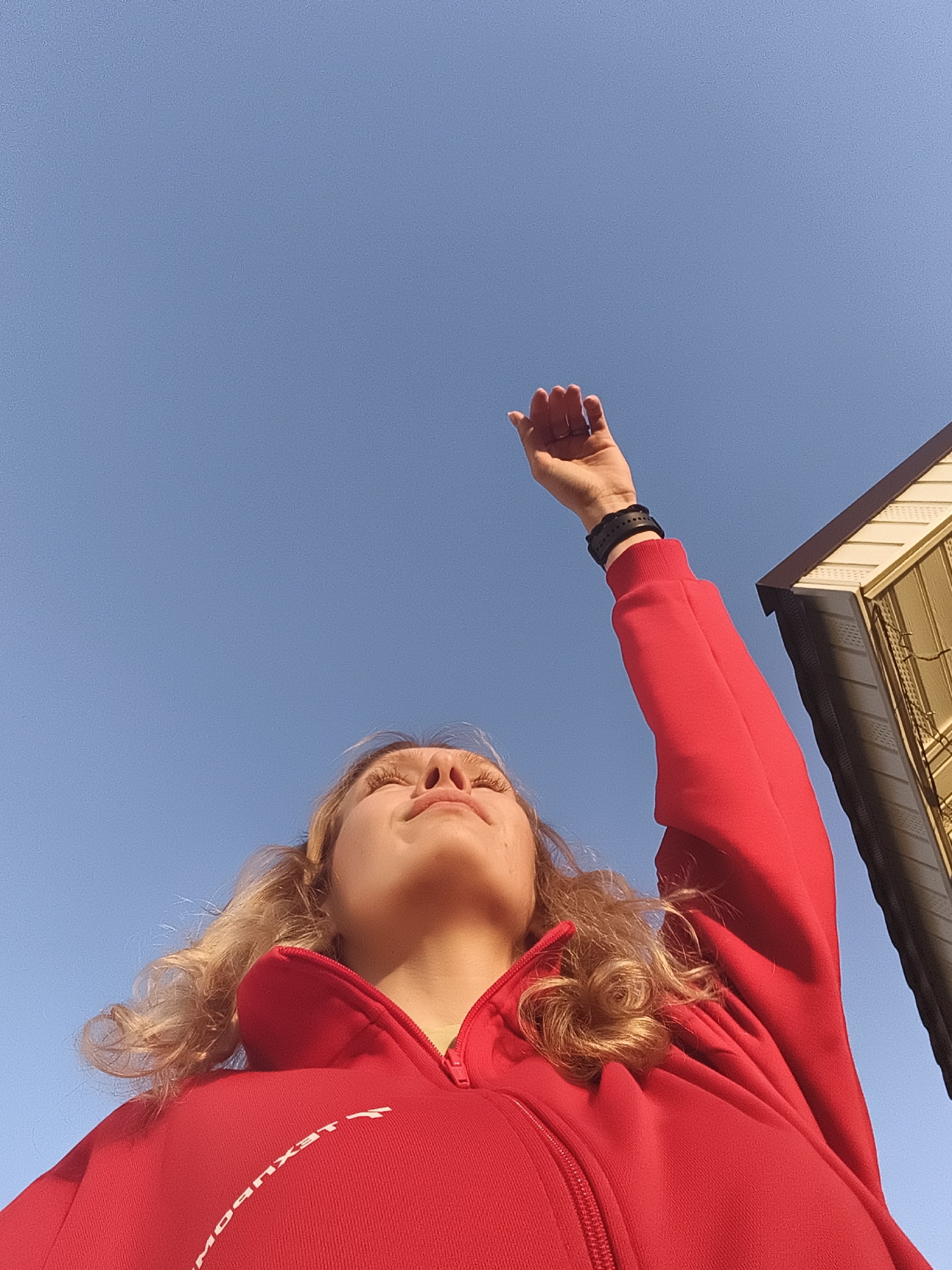 The image depicts a person in Русский Вожой, Russia, wearing a red zip-up jacket, with their arm raised towards the sky. The sky is clear and blue. Part of a building is visible in the background. The person is looking upwards, their eyes are closed. They have blonde curly hair. A black bracelet or watch band is visible on their raised wrist. The jacket has white lettering visible at the bottom corner, reading "LEXUBOM".