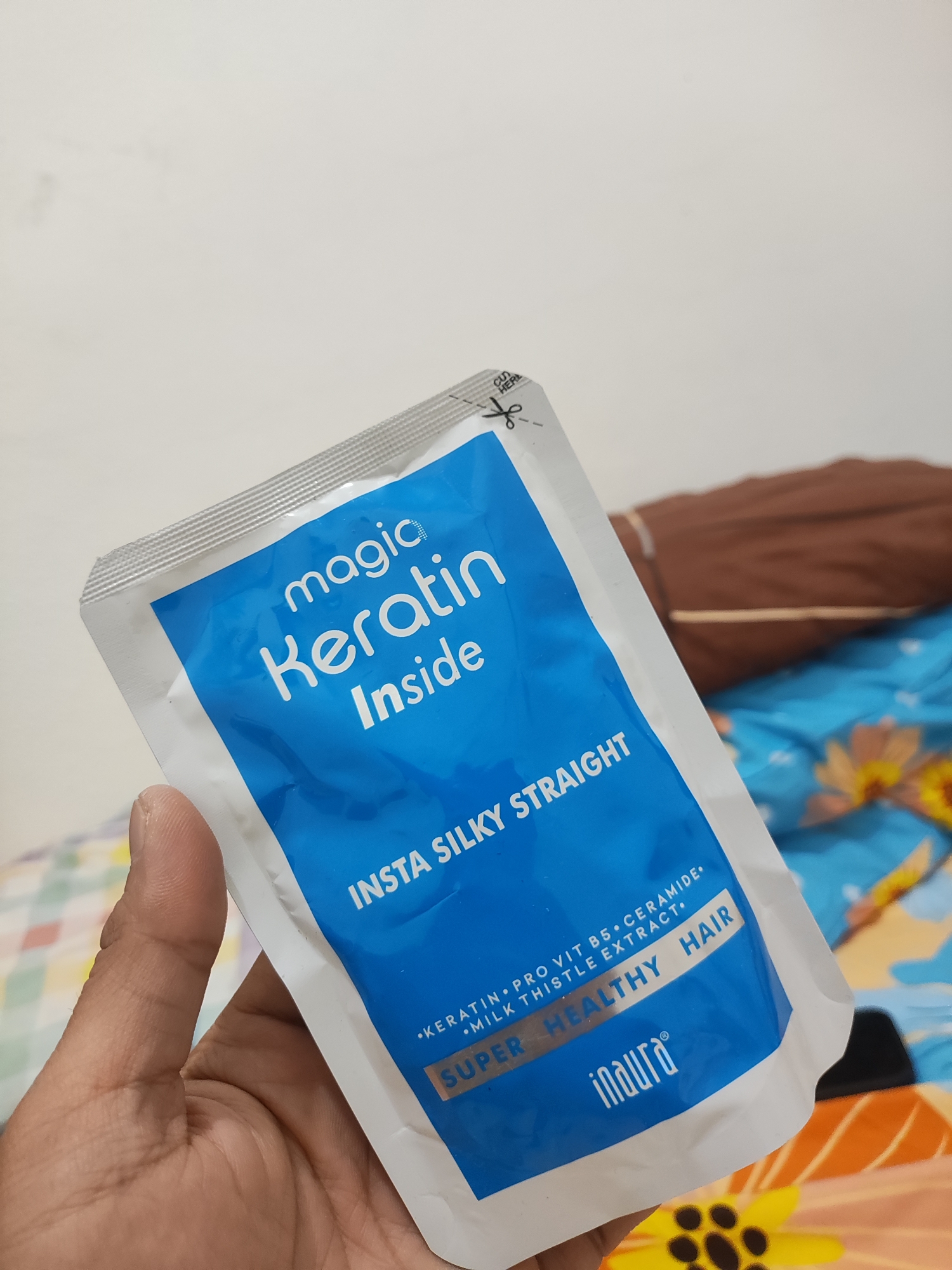A close-up, eye-level shot shows a person's hand holding a small, blue and white pouch of "magic Keratin Inside" hair product. The pouch has silver, serrated edges and a "cut here" indicator with scissors printed on it. Text on the pouch reads "INSTA SILKY STRAIGHT," and smaller text lists ingredients. The Inaura logo is visible at the bottom. The background is out of focus, showing a patterned blue and yellow cloth, possibly bedding, and a brown pillow. The image was taken in Wanajaya, Indonesia.