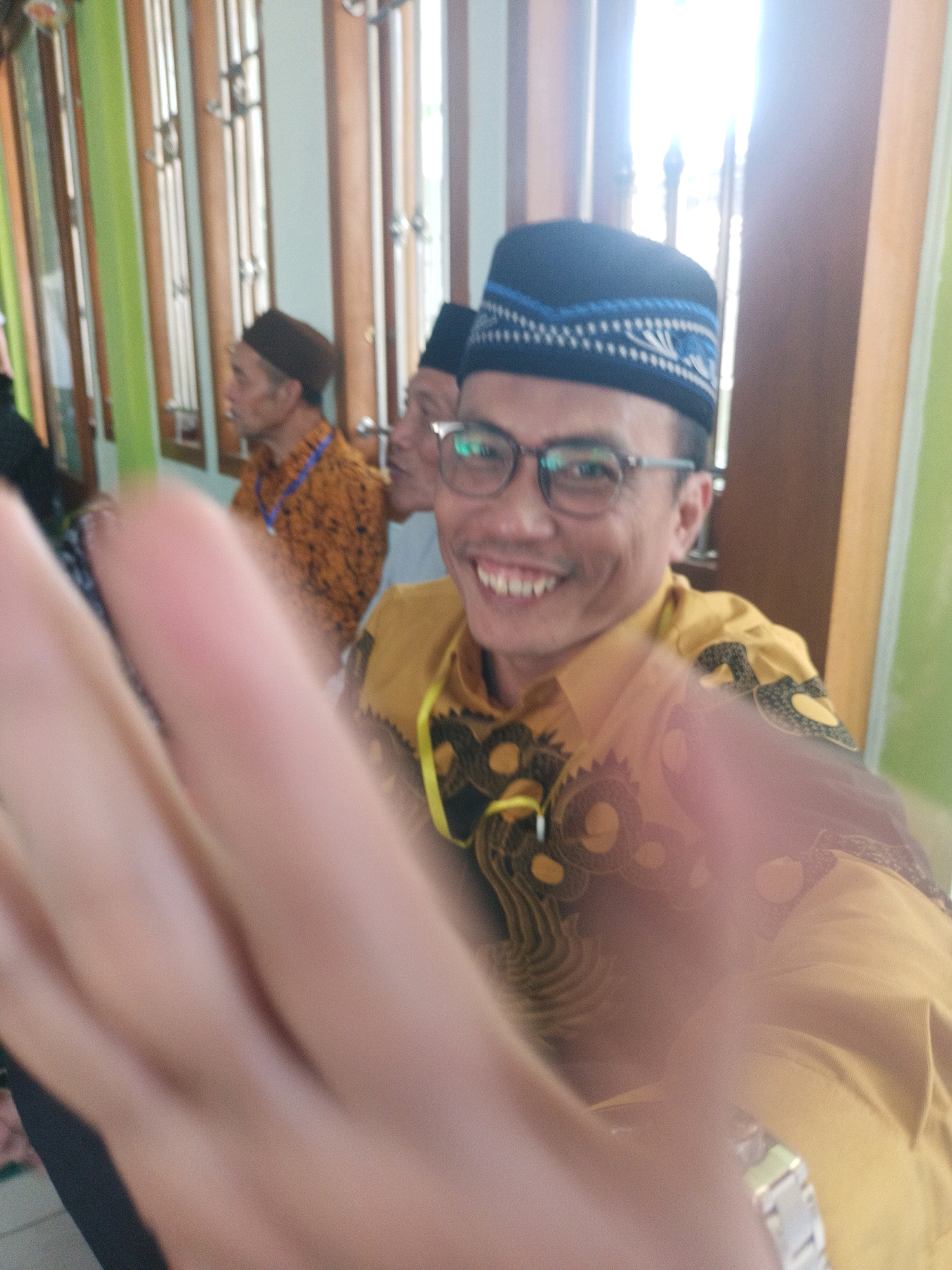 In Penusupan, Indonesia, a man wearing a black and blue patterned peci cap, glasses, and a yellow batik shirt with a yellow lanyard smiles at the camera. He is in focus, with a blurred hand in the foreground, suggesting the photo was taken by himself. Behind him, two other men are partially visible. One man in the middle ground wears an orange batik shirt and a brown peci cap. Another man is visible to his left. The background consists of wooden window frames and green walls.