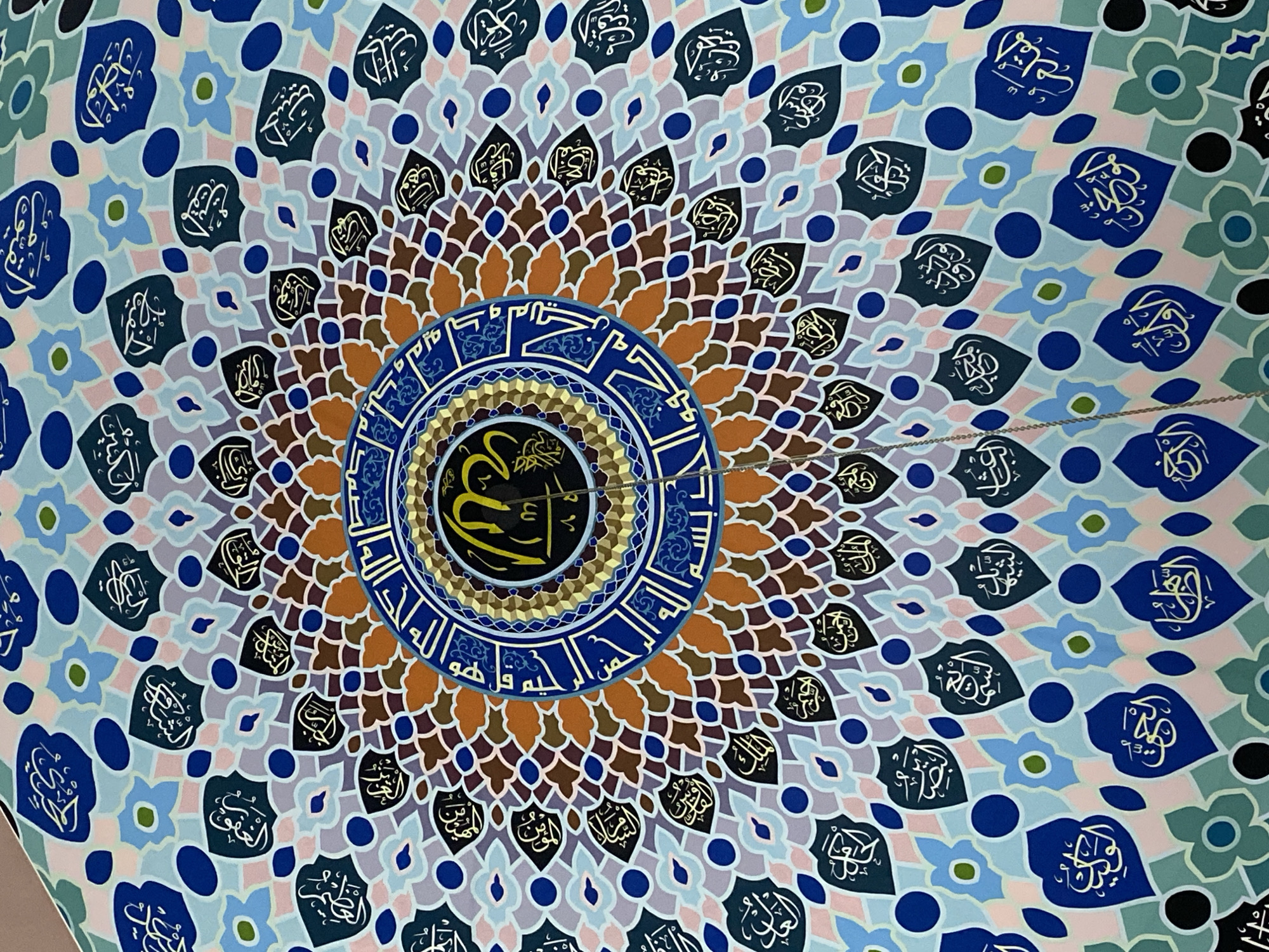 This is a medium shot, eye-level, indoor photograph of an ornate ceiling with Islamic calligraphy. The calligraphy is arranged in a circular, mandala-like pattern with intricate geometric designs and floral motifs. The colors are predominantly blues, purples, browns, oranges, and light pinks, with white and gold calligraphy. The overall effect is one of grandeur and religious devotion. The photograph seems to have been taken in a place of worship or a building with significant cultural or religious importance. The text visible in the image consists of Arabic script, likely verses from the Quran or religious phrases. Based on the location context provided, this ceiling is likely located in Lhokseumawe, Indonesia. The lighting appears to be artificial and even, suggesting an indoor setting at any time of day. There are no people or objects other than the ceiling itself visible in the frame. The style of the calligraphy and the geometric patterns are typical of Islamic art and architecture.