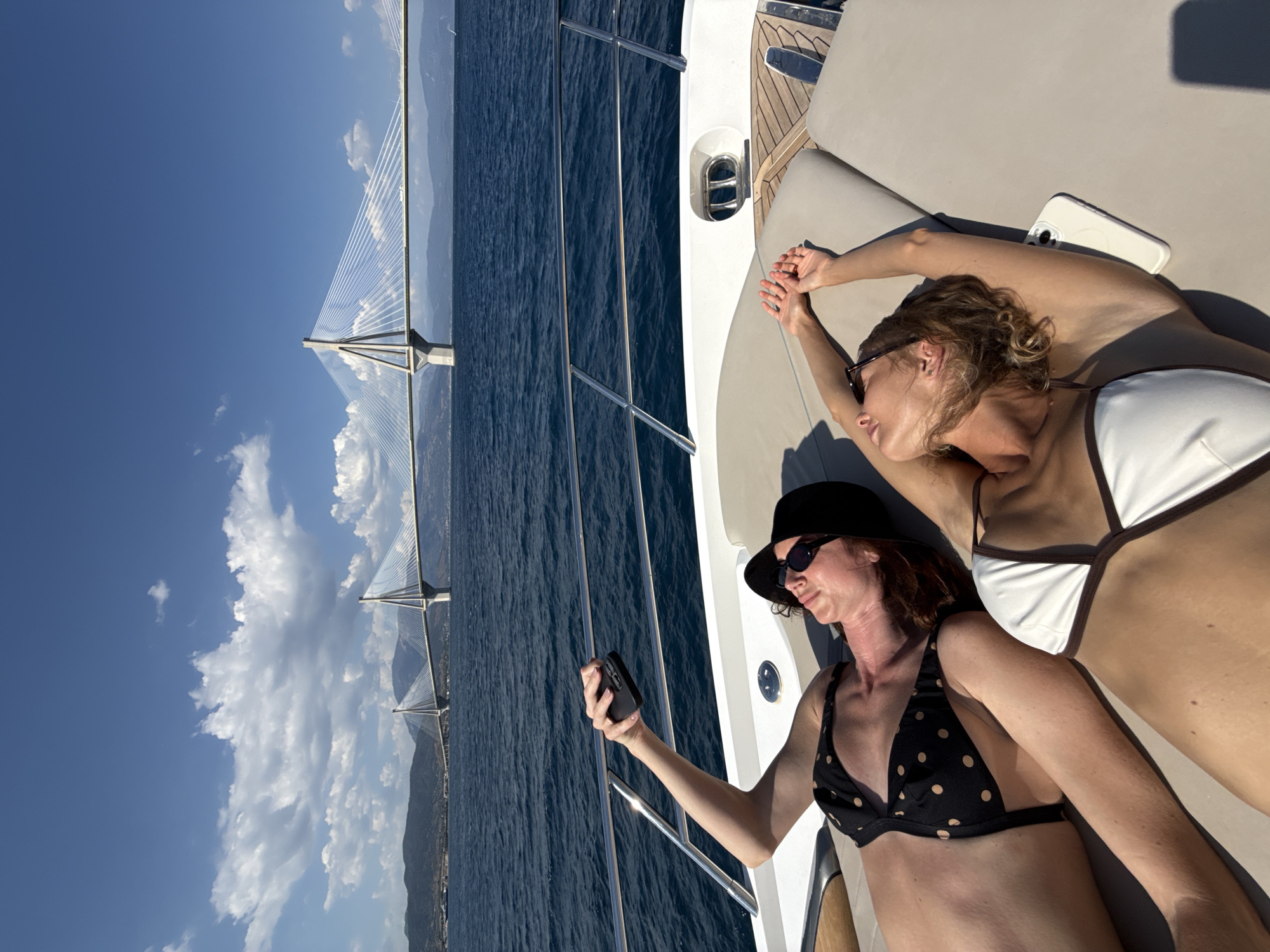 The media file shows two women relaxing on the deck of a yacht. The woman in the foreground is wearing a black polka-dot bikini and a black hat, and is looking at her phone. The woman in the background is wearing a white bikini and has her arms raised above her head. The yacht is on the sea, with a large cable-stayed bridge visible in the distance. The sky is blue with some white clouds, suggesting a sunny day. The scene appears to be set in the Municipal Unit of Rio, Greece, as indicated by the prominent bridge which is part of the Rio–Antirrio Bridge. The women appear relaxed and are enjoying their time on the water. There is no visible text in the media file.