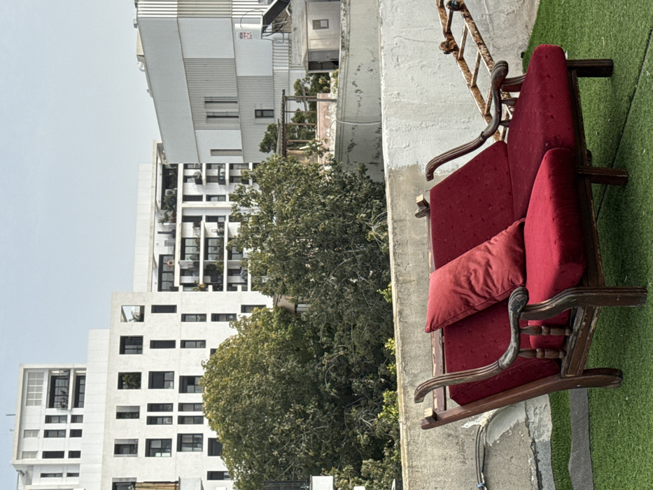 A rooftop scene in Tel-Aviv, Israel, captured under a pale blue sky. In the immediate foreground, a two-seat antique-style wooden daybed or bench, featuring dark red velvet upholstery and one matching red rectangular pillow, rests on artificial green turf. The wooden frame of the seating exhibits visible wear and weathering. Behind the seating is a low, textured concrete parapet wall, painted light gray/white, with some paint degradation. Beyond the parapet, a midground area contains a dense cluster of green trees. Above and behind the trees, various urban rooftop structures are visible, including a small, light-colored corrugated metal utility shed and a wooden pergola structure. The background is dominated by several modern, multi-story residential buildings with stark white facades, numerous dark-framed windows, and multiple balconies. No individuals are present within the frame, and no discernible actions or interactions are depicted.