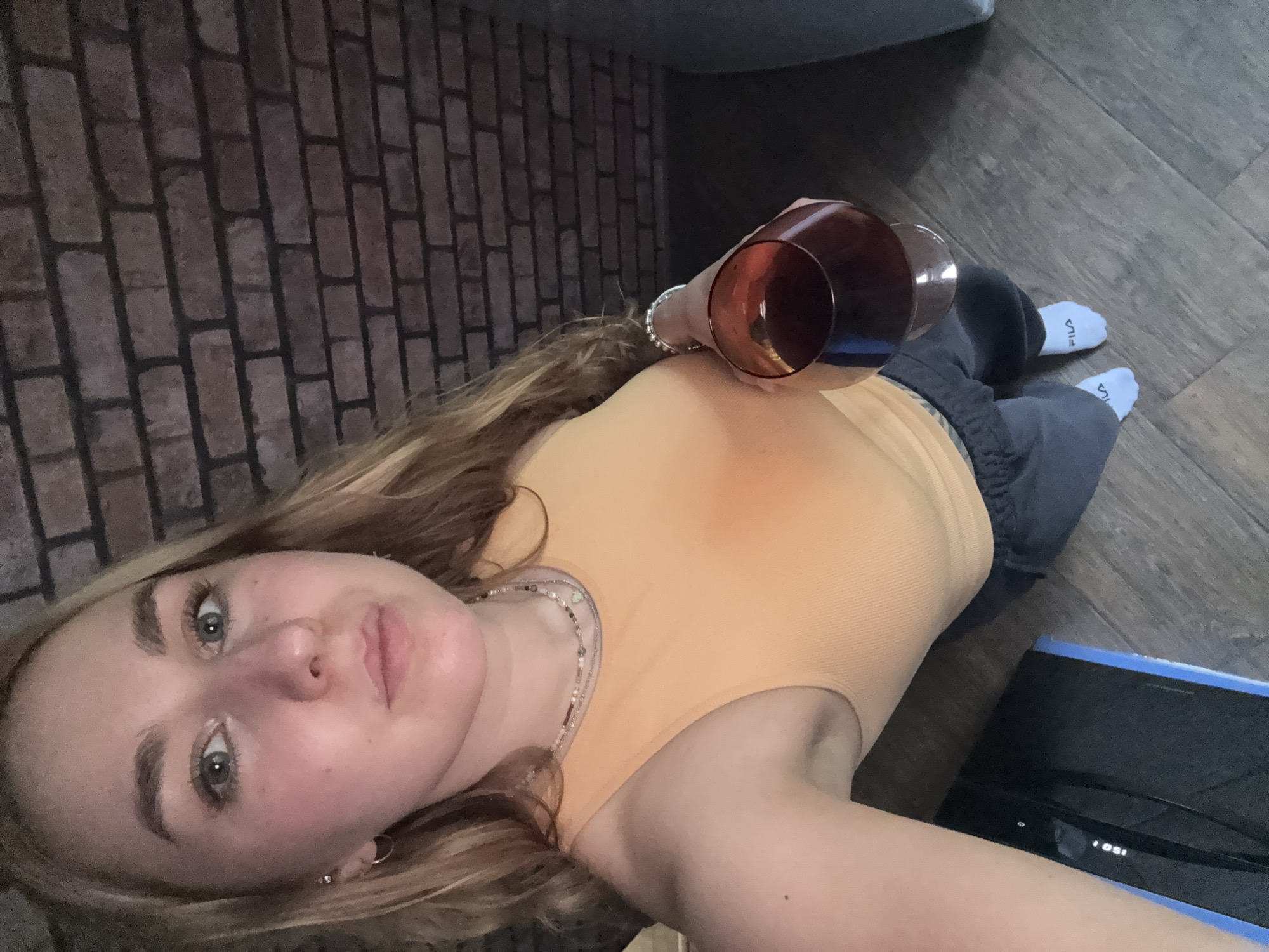A young woman with blonde hair is lying on a wooden floor, holding a glass of red wine. She is wearing an orange tank top, grey sweatpants, and white socks with a "FILA" logo. A blue-edged, black electronic device sits to her right. A brick wall is visible behind her. The location cannot be determined from the image.